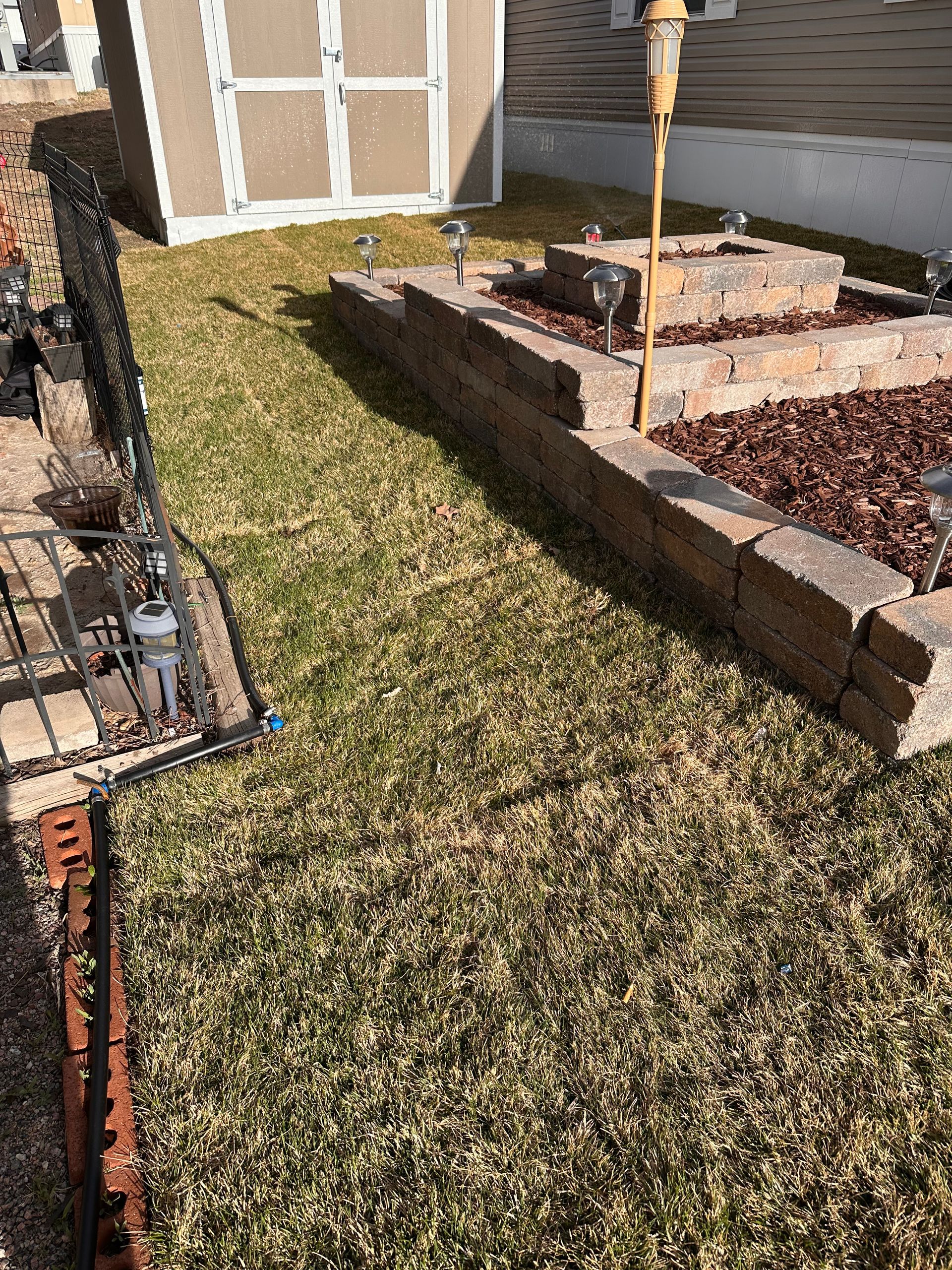 Backyard garden with a raised bed, grass, and a trellis near a brick wall