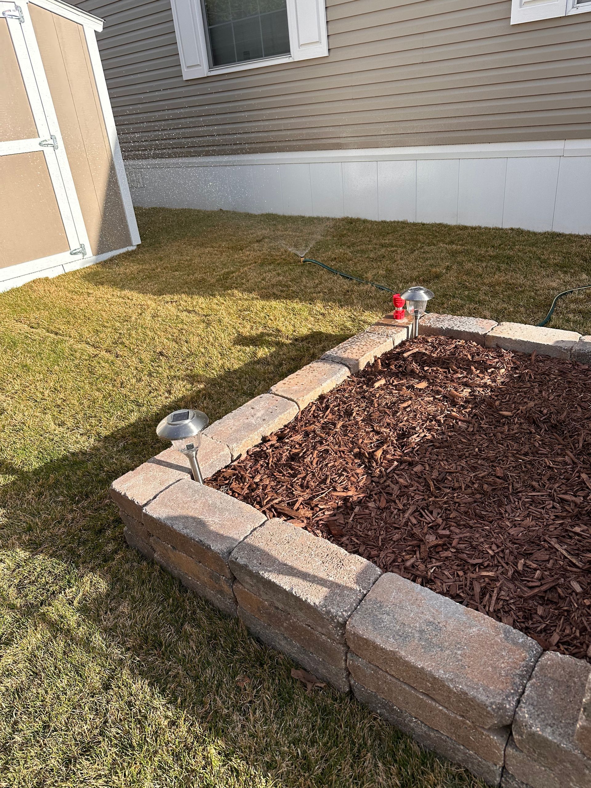 Backyard garden bed with stone border, brown mulch, grass, and a small red sprinkler near a house wall