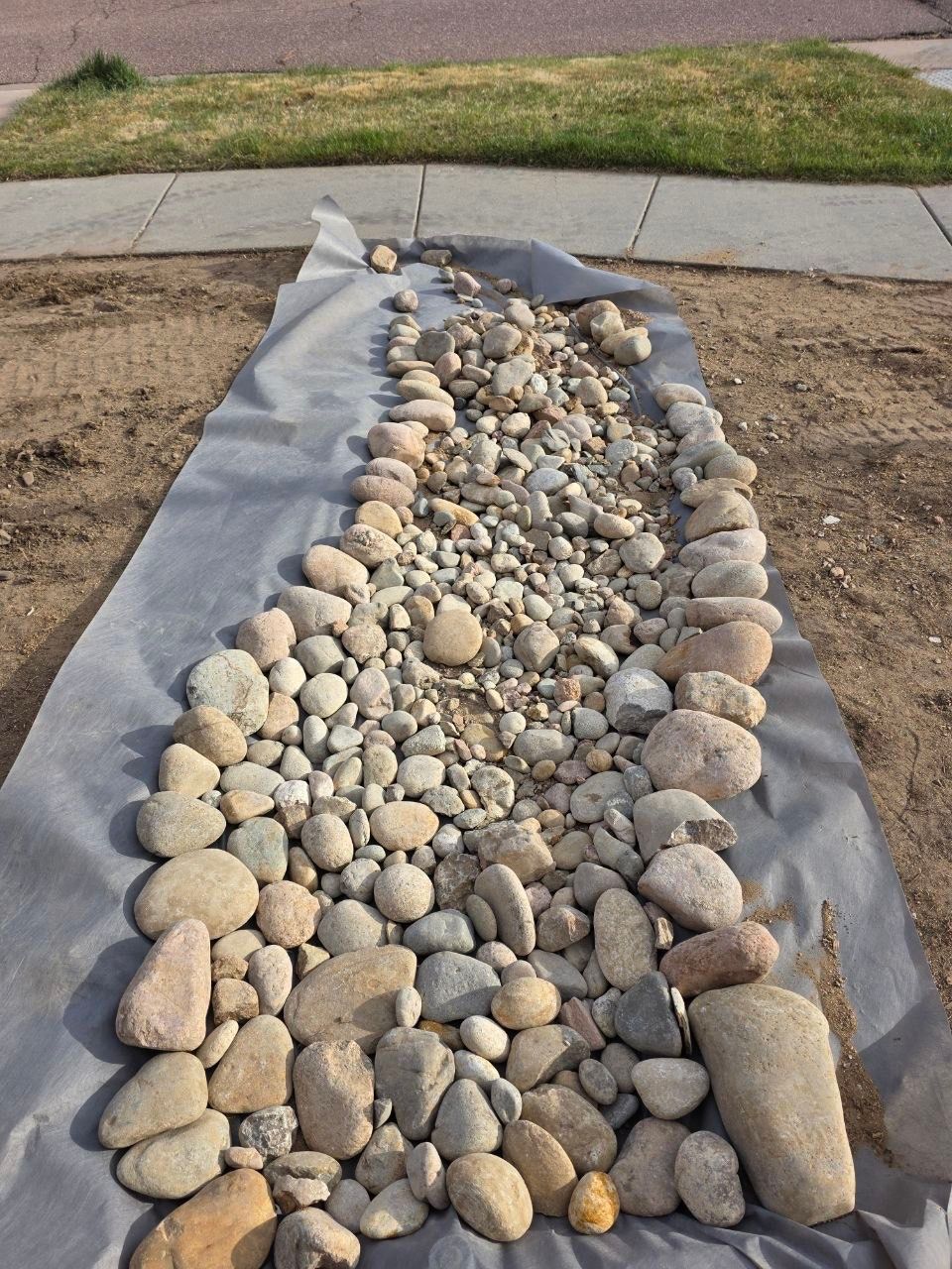 Gray landscape fabric covered with assorted river rocks in a garden bed beside a sidewalk