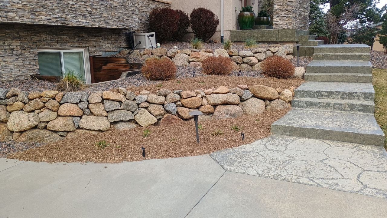 Stone retaining wall with desert landscaping and concrete steps leading up to a stone house entrance