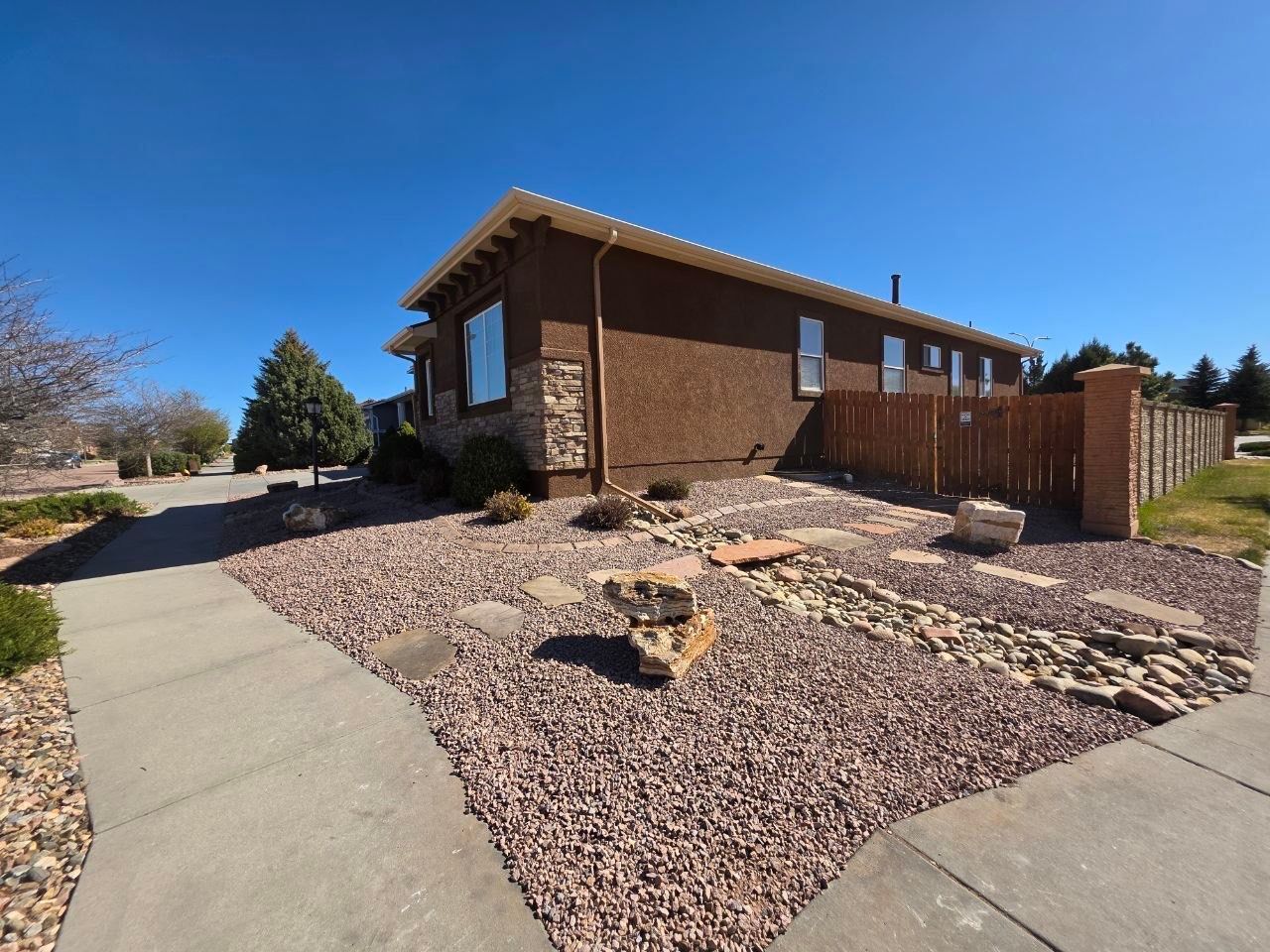 Single-story brick house with a sloped rock garden, concrete walkway, and wooden fence under a clear blue sky
