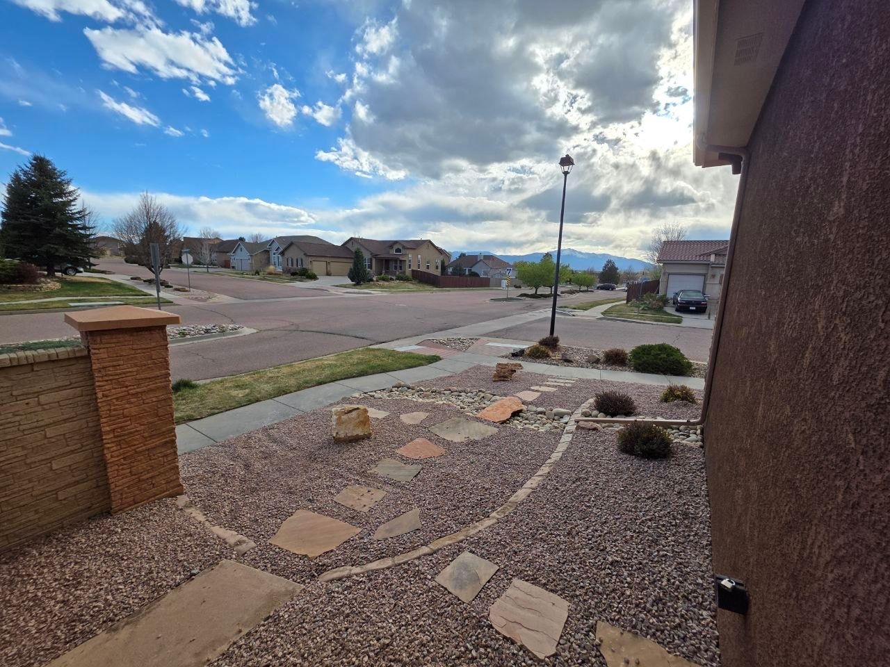 Suburban front yard with stone path, brick wall, driveway, cloudy sky, and houses in the distance