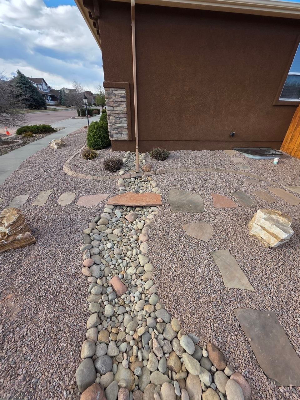Desert-style front yard with a pebble path, stepping stones, and a brown stucco house under a cloudy sky