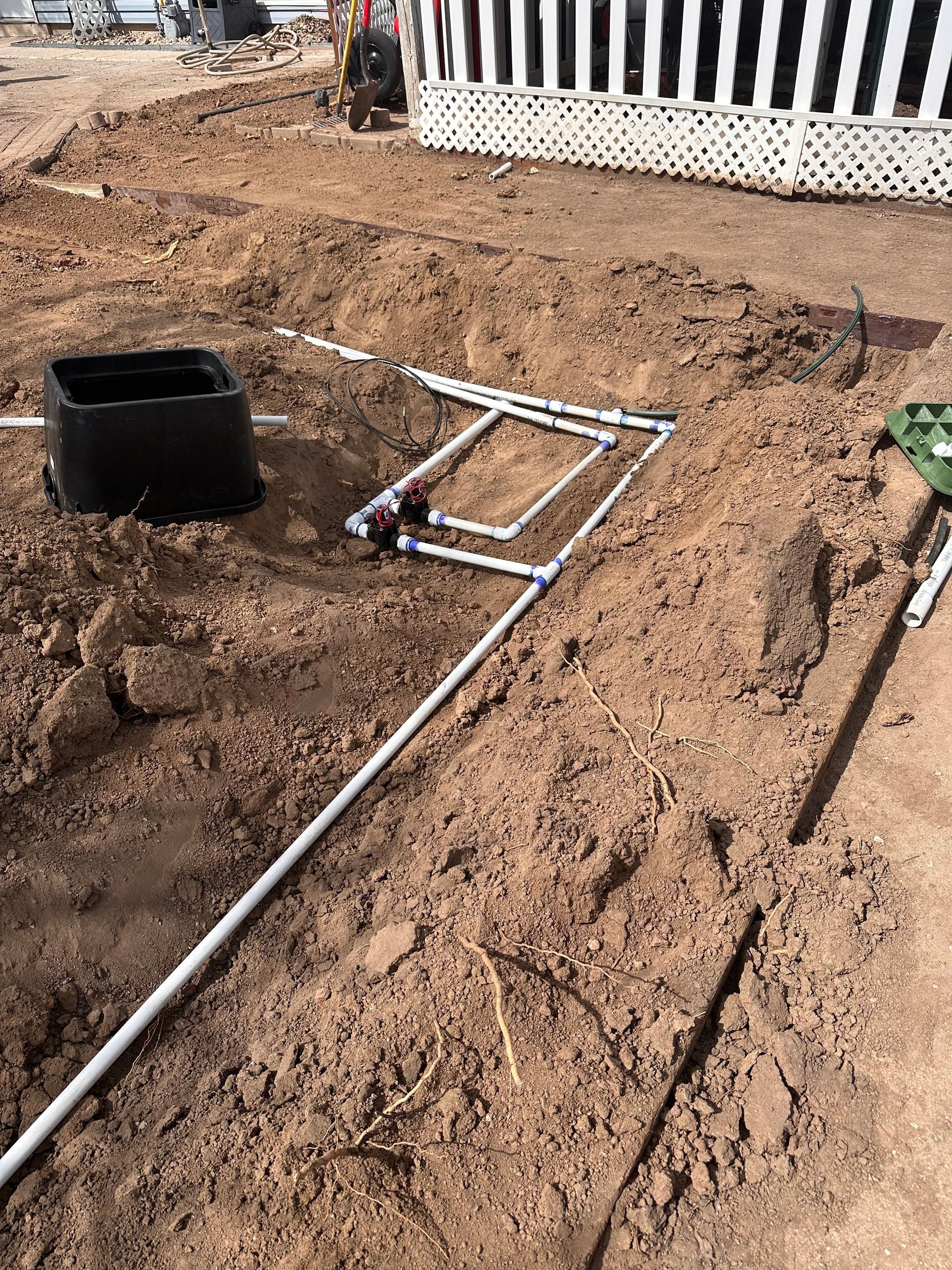 Trench and PVC piping in a dirt yard with a black bucket and a white lattice fence in the background
