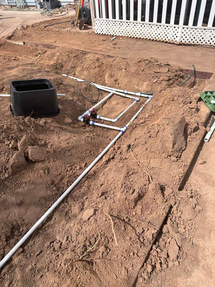 White PVC pipes and valves laid in a trench, near a black box and a building, in dirt.