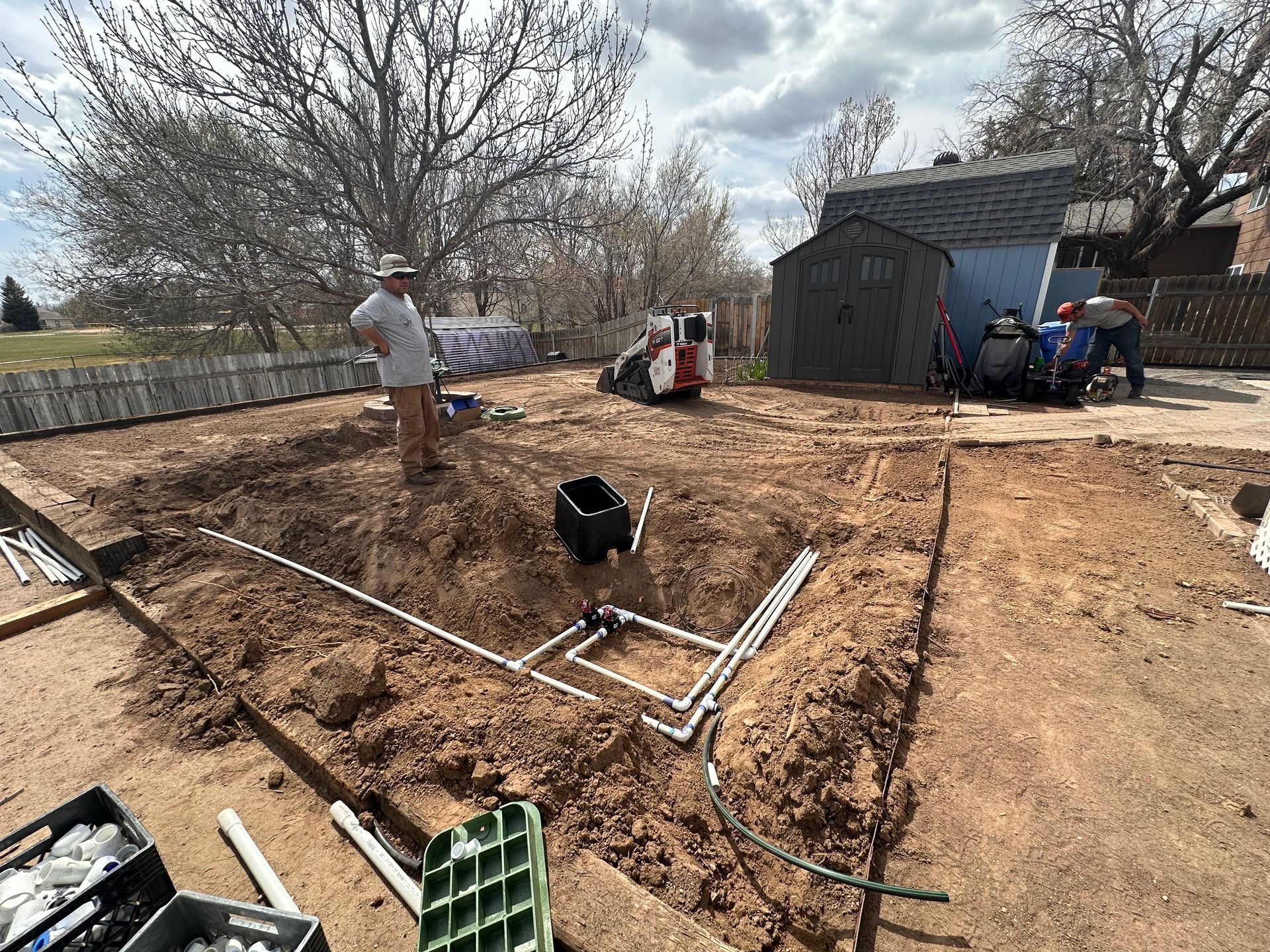 Backyard excavation with pipes and tools, workers near a shed under a cloudy sky