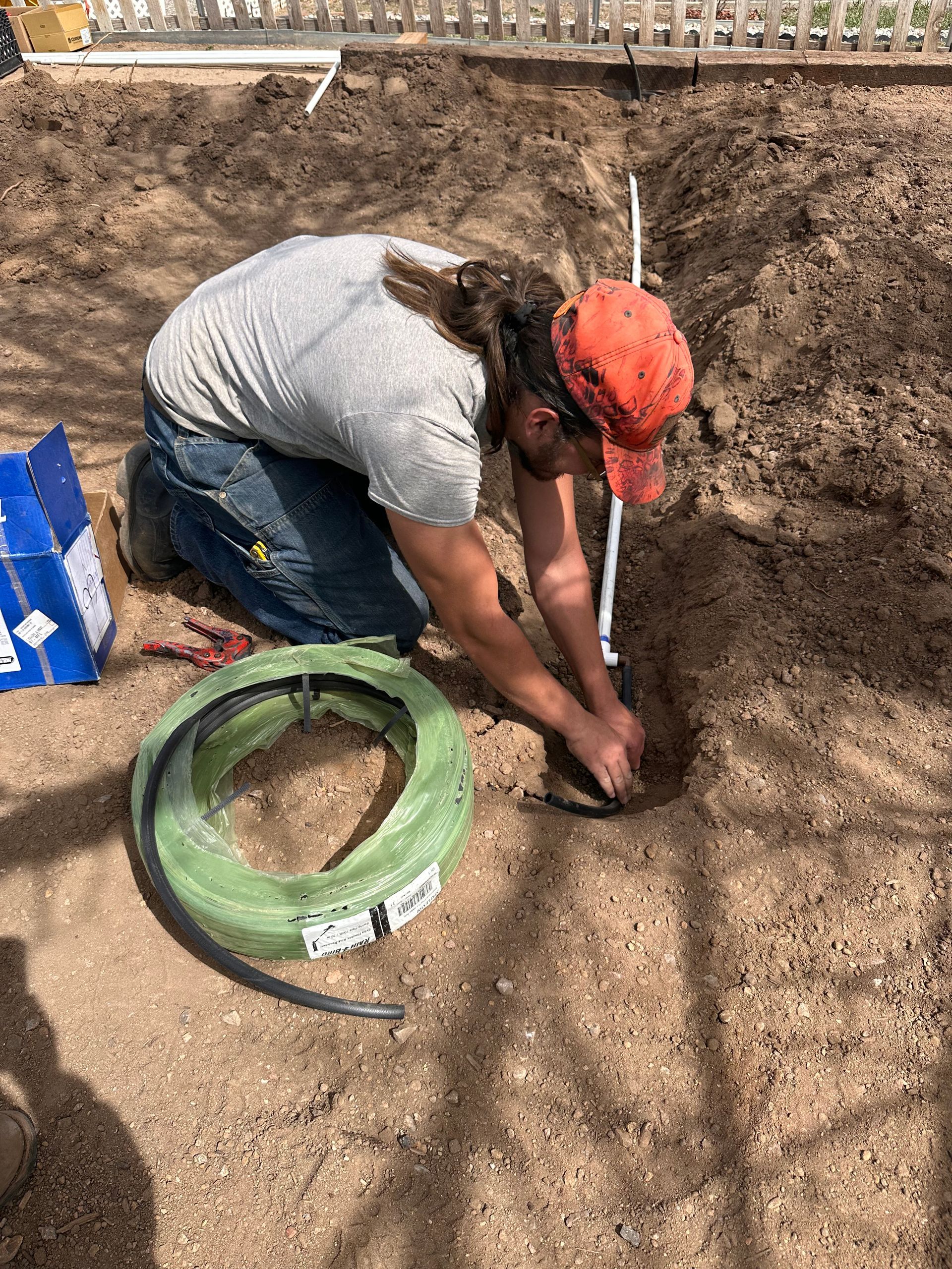 Worker kneeling in a dirt trench, digging with a hand tool beside a green bucket and blue tarp