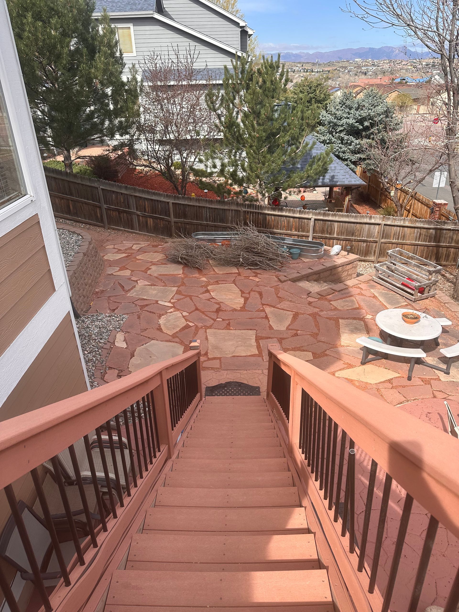 View down wooden stairs to a stone patio, trees, and a city view beyond a hillside backyard.