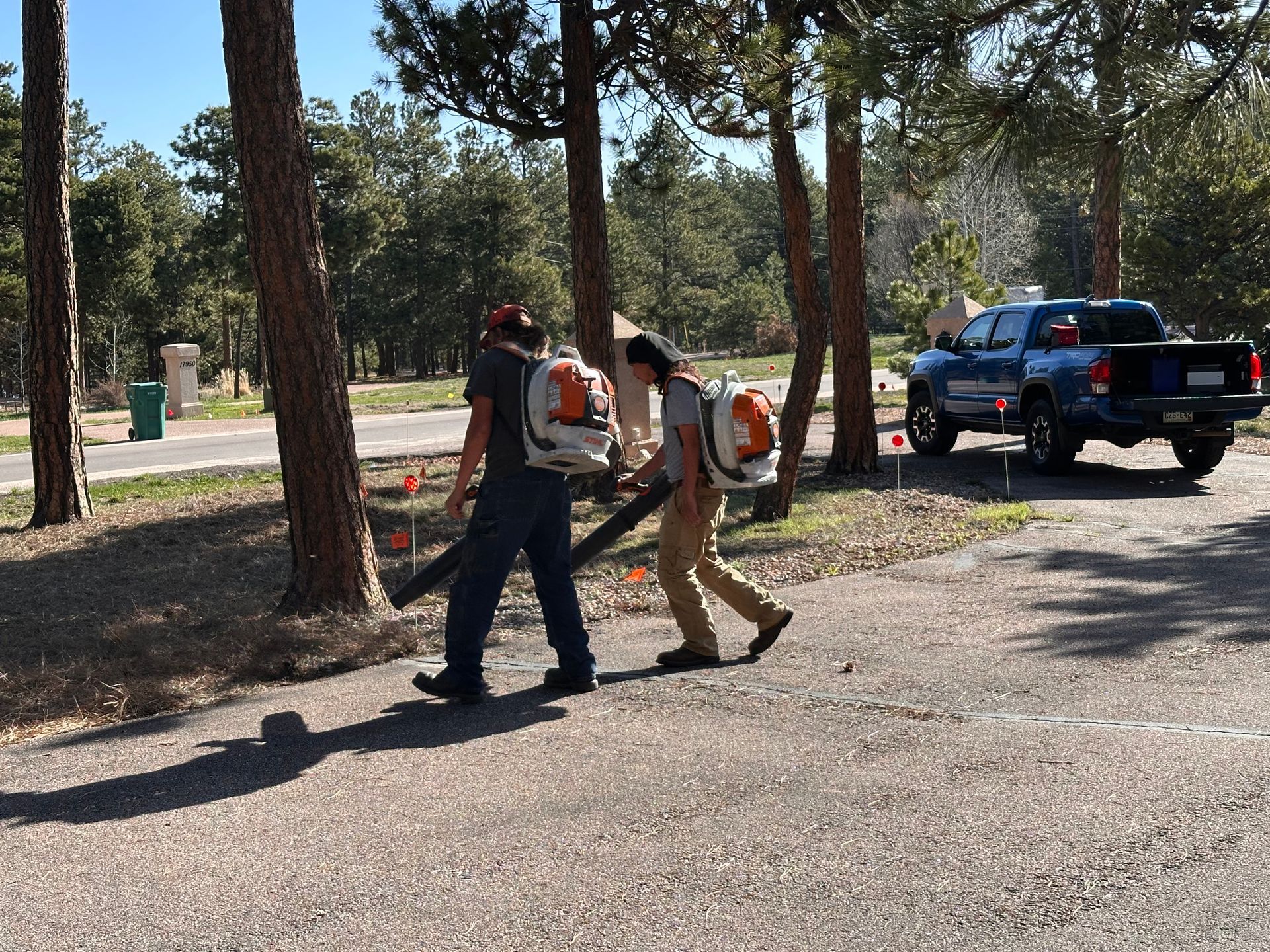 Two workers walking with tools near a blue pickup truck in a wooded roadside area.