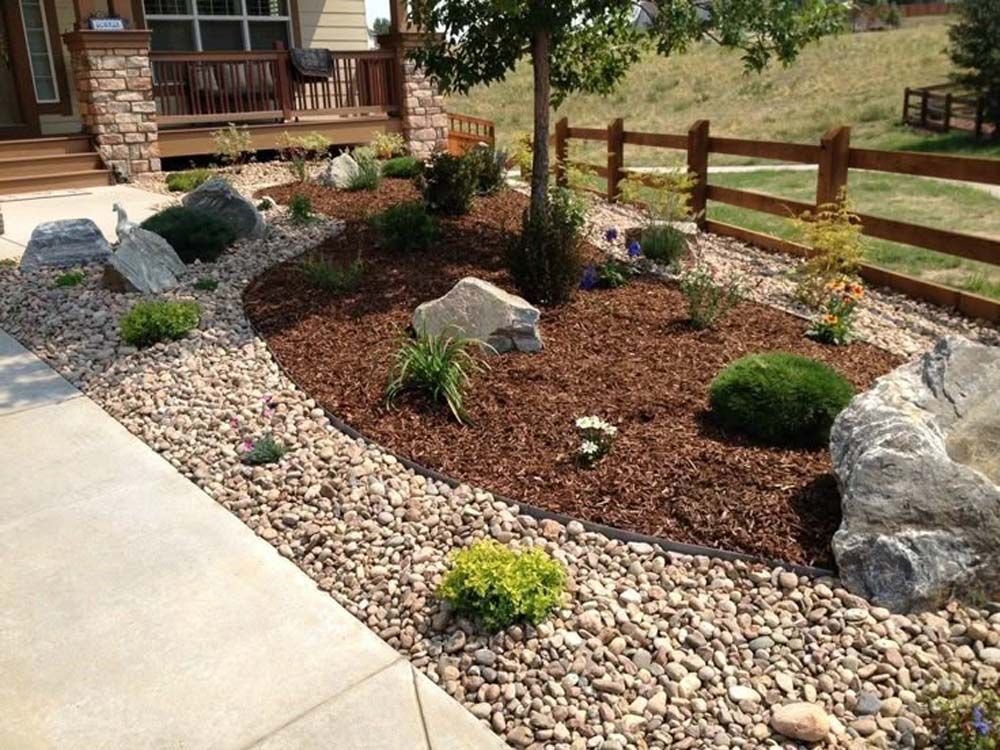 Landscaped front yard with rocks, mulch, shrubs, and a tree along a sidewalk and fence.