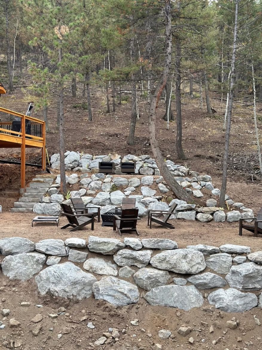Stone fire pit and seating area on a wooded hillside with a wooden deck and retaining wall