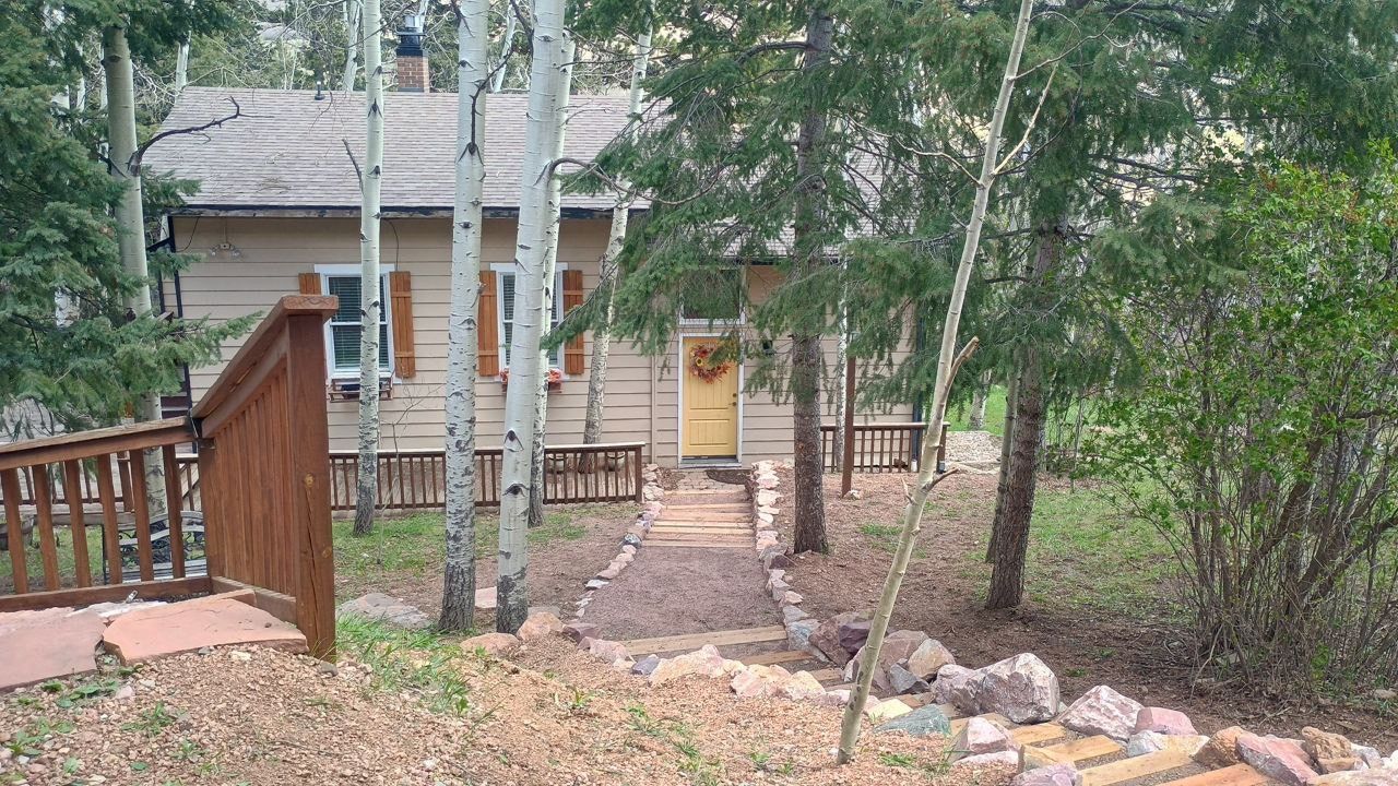 Wooden house with a front porch, stone walkway, and trees in a wooded yard.