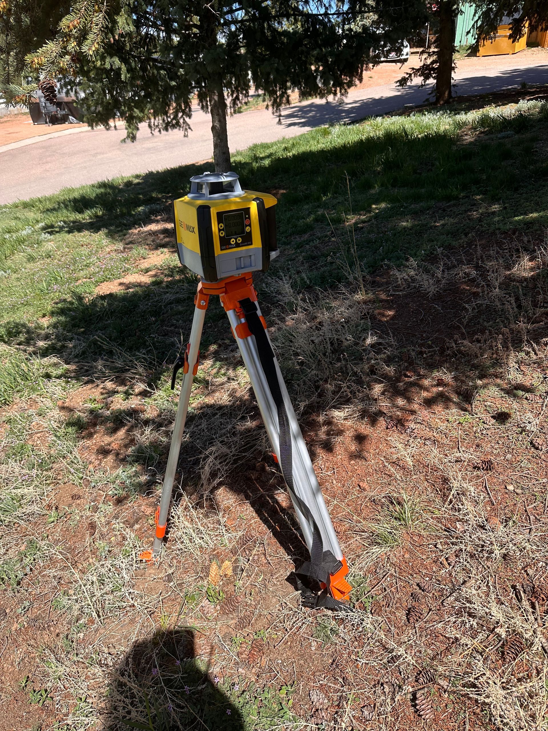 Yellow surveying instrument on a tripod in a grassy, sunlit outdoor area under trees.