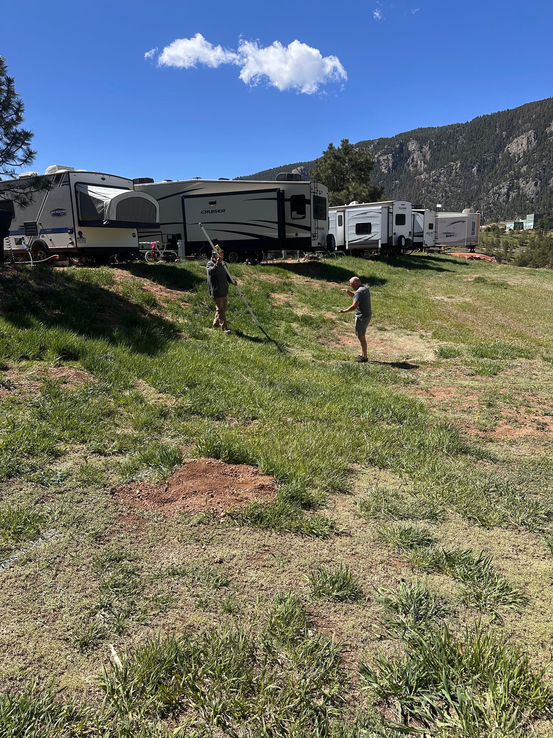 RVs parked on a grassy hillside with two people walking under a blue sky and mountains in the background