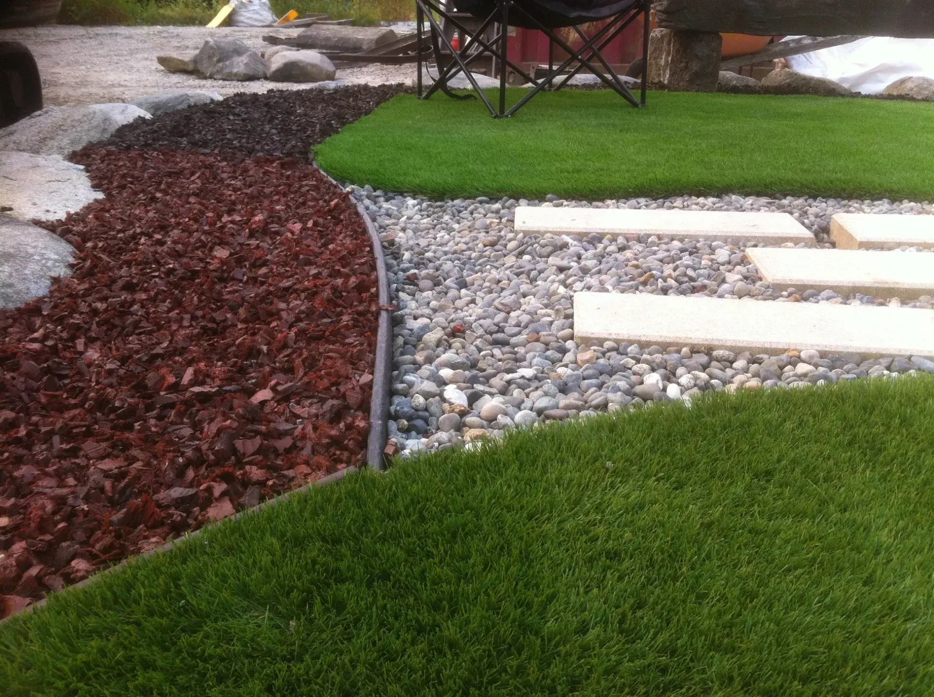 Curved garden bed with red mulch, gray stones, and green grass beside a patio and chair