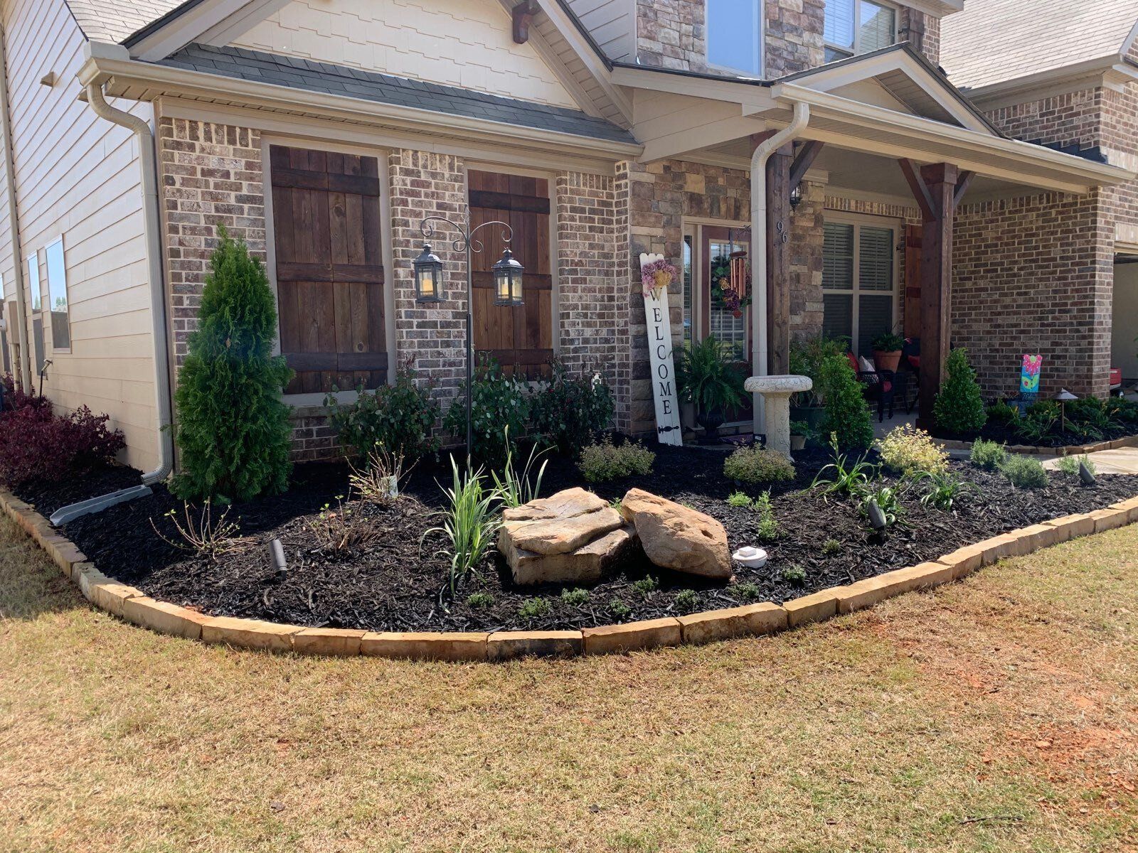 Brick house front yard with mulched flower bed, small shrubs, large rocks, and a birdbath