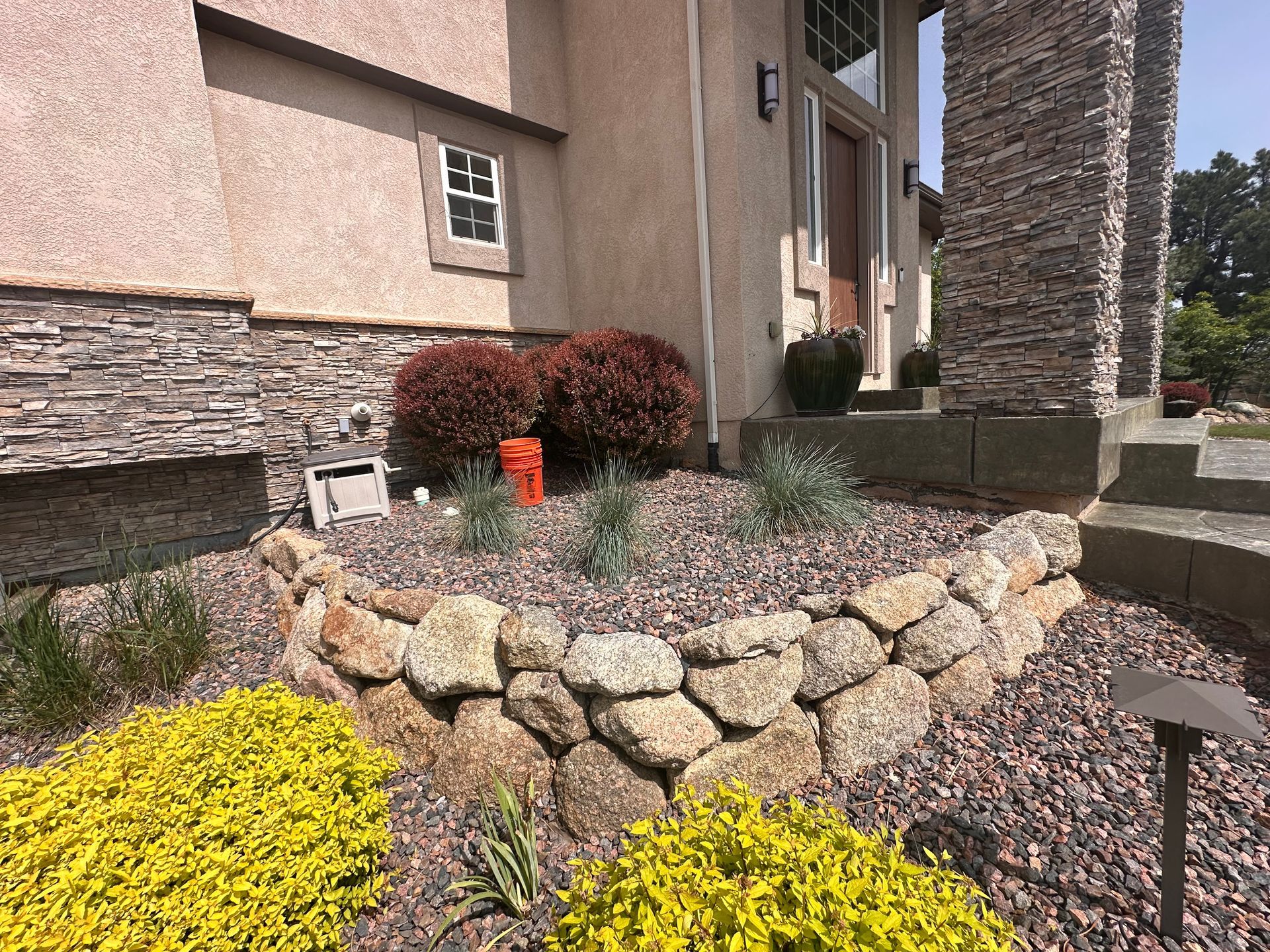 Landscaped front entrance with stone retaining wall, shrubs, and yellow flowers beside a house