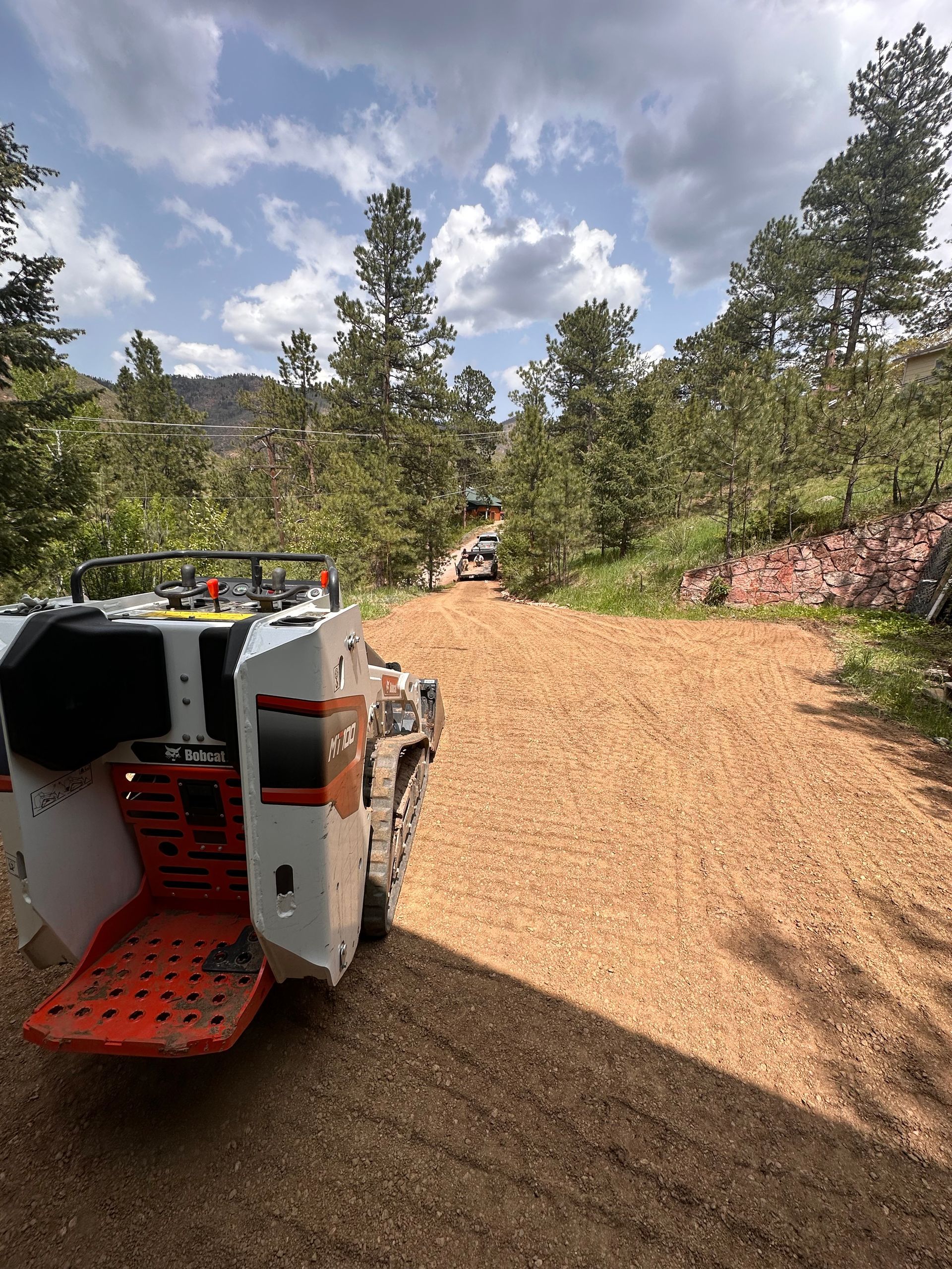 White off-road vehicle on a gravel mountain road with pine trees and cloudy sky.