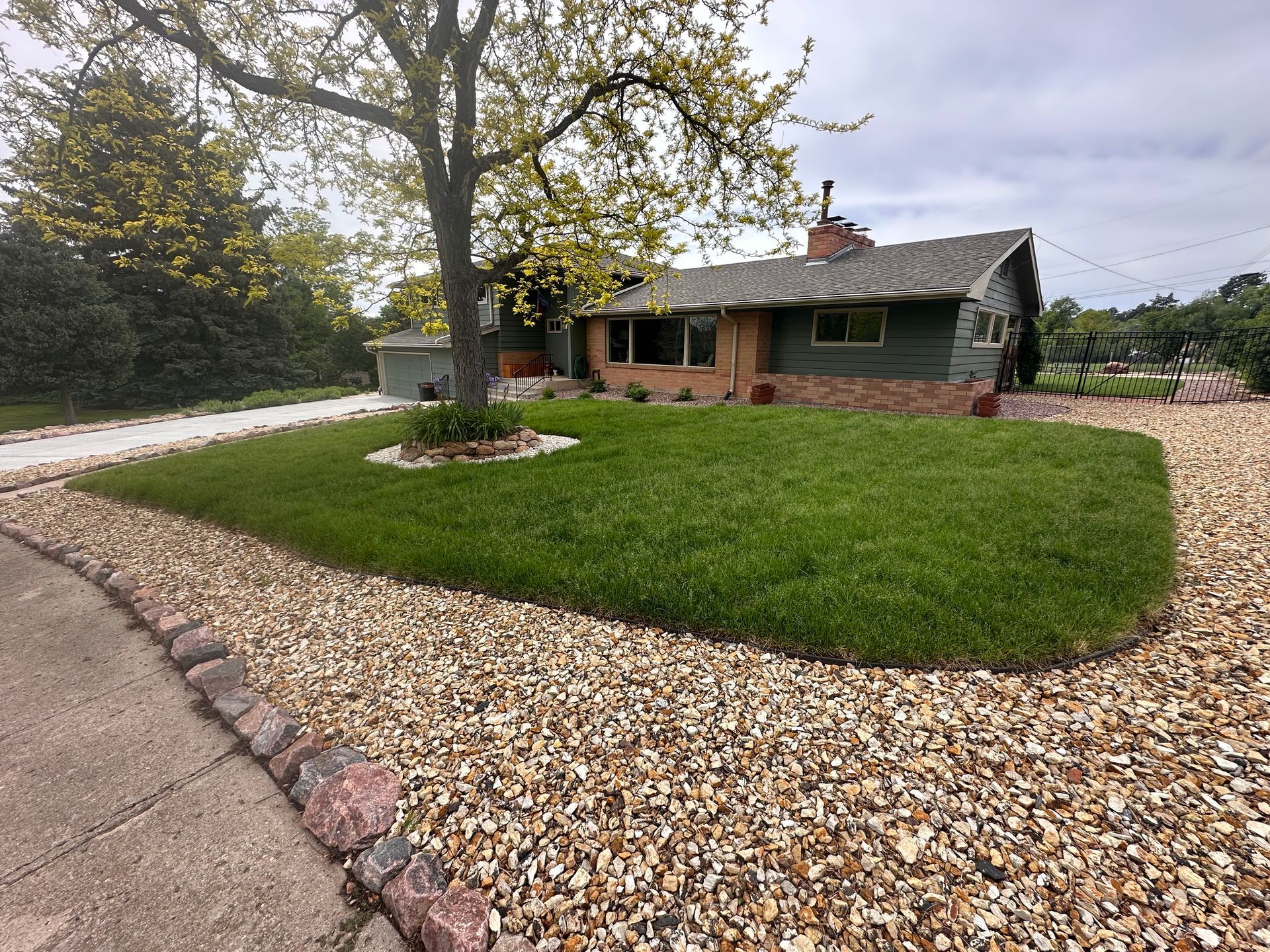Single-story brick house with a large green lawn, gravel border, and a tree in the front yard.