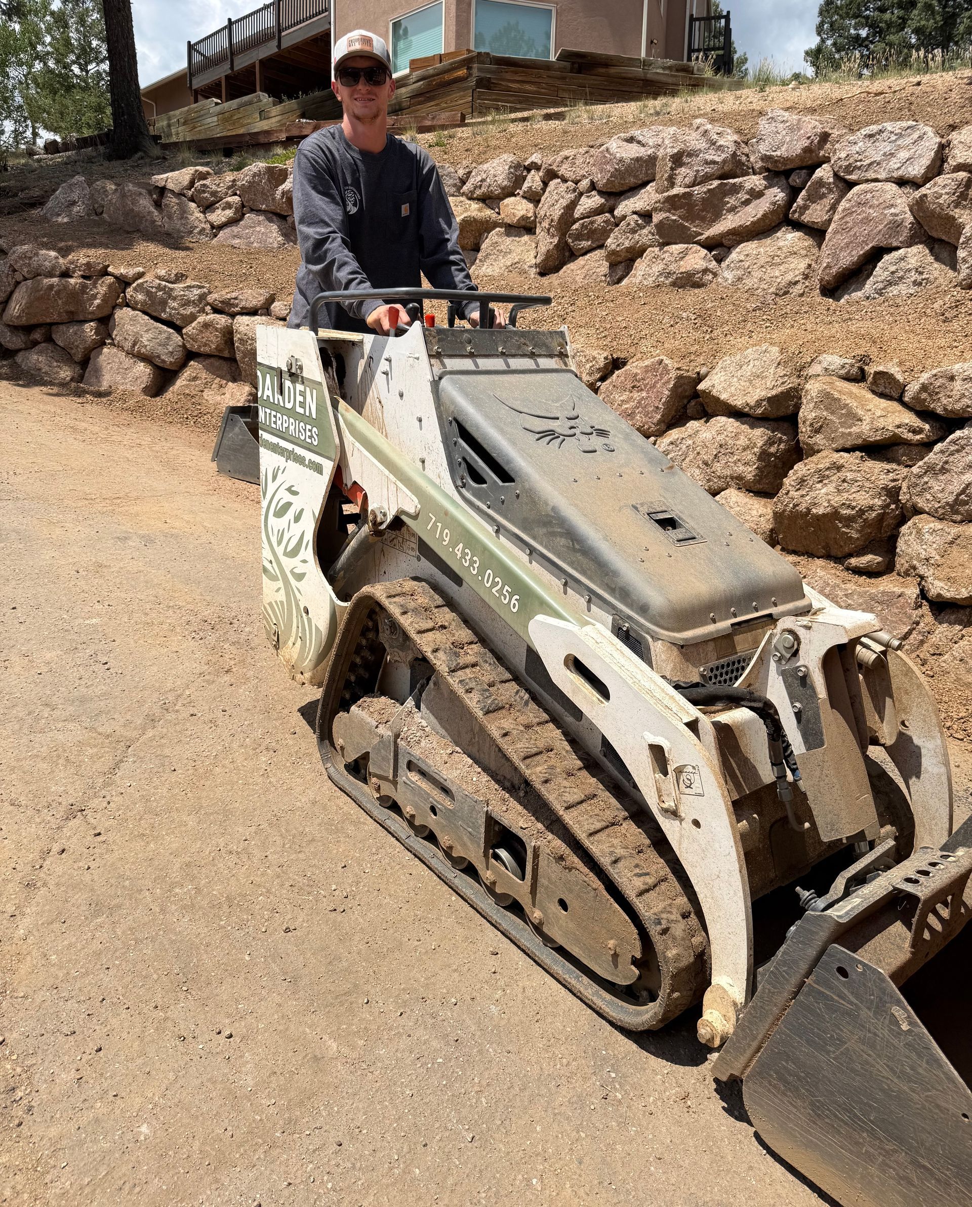 Person operating a small tracked construction machine on a gravel path beside a stone wall.