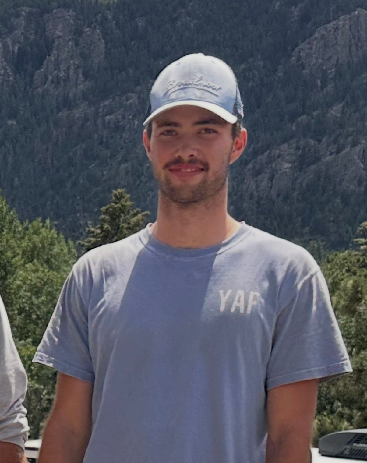 Young man in a light blue cap and shirt standing outdoors with mountains behind him