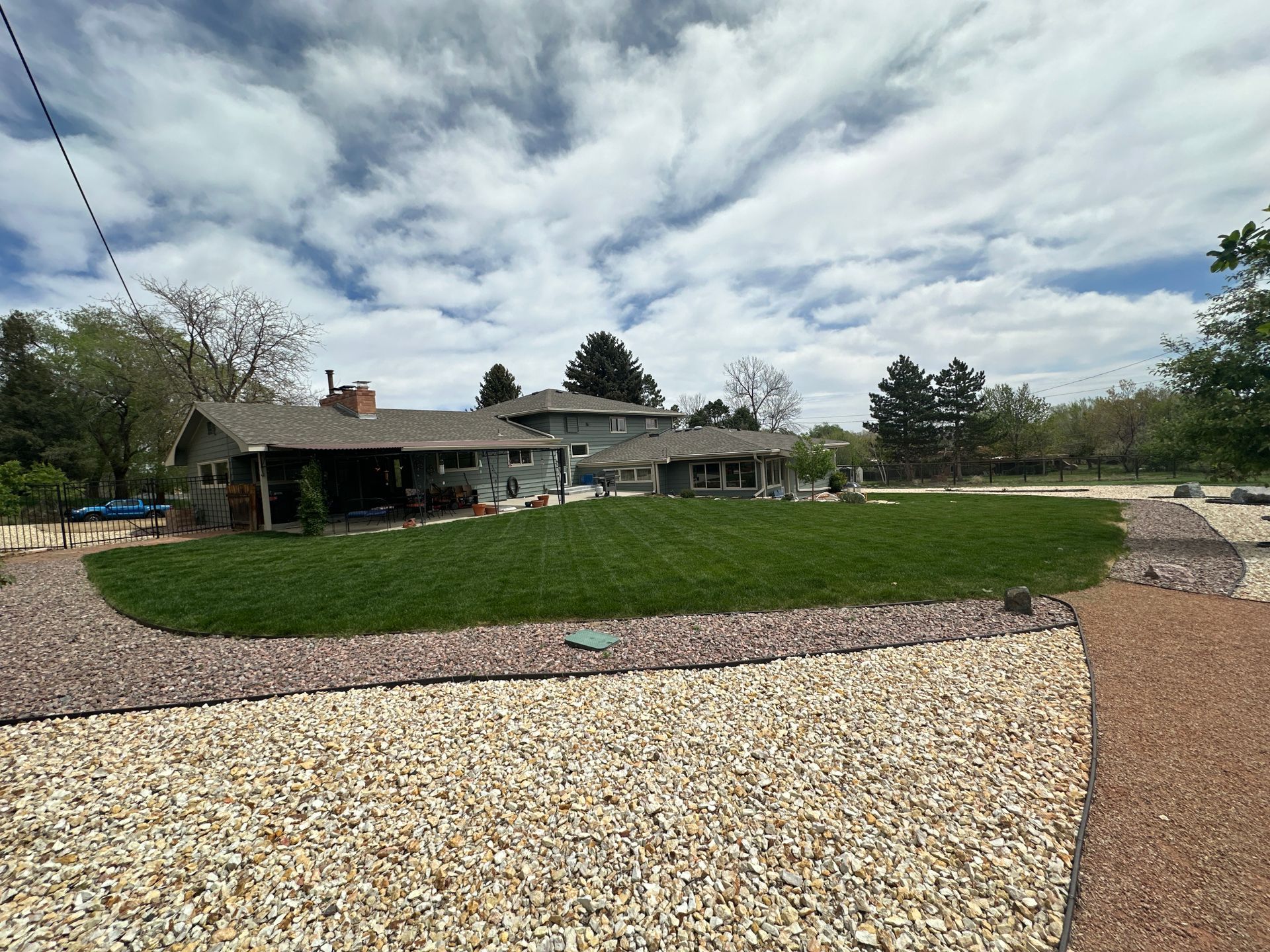 Residential backyard with a large green lawn, gravel paths, and a cloudy sky above a house.