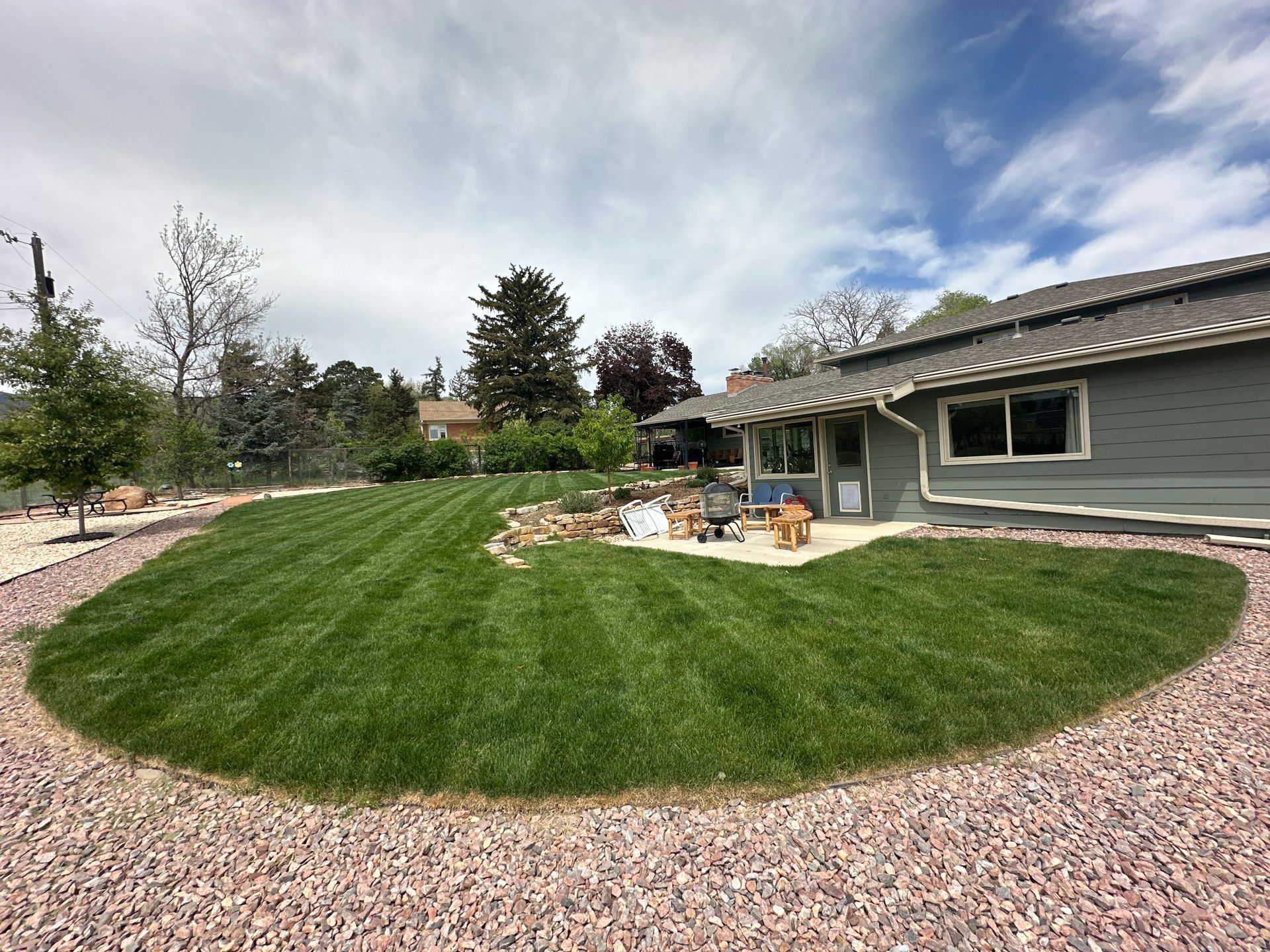 Backyard patio beside a gray house with green lawn, gravel border, and patio furniture under a cloudy sky