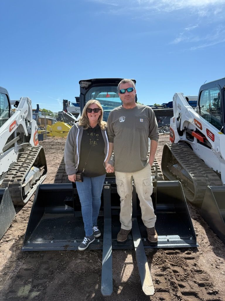 Two people standing in front of construction equipment under a clear blue sky