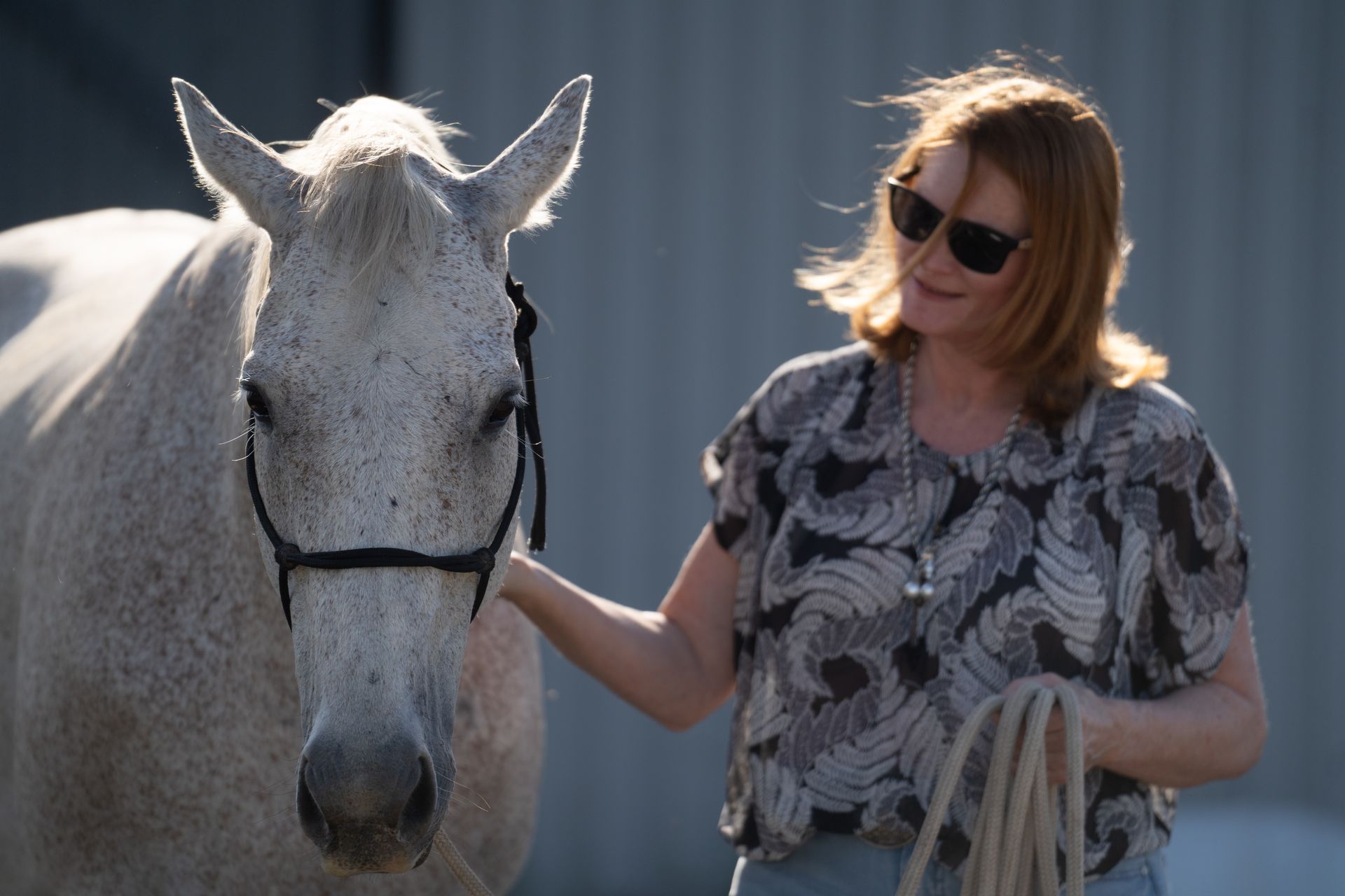 Woman in sunglasses with a gray horse outdoors, touching its neck.