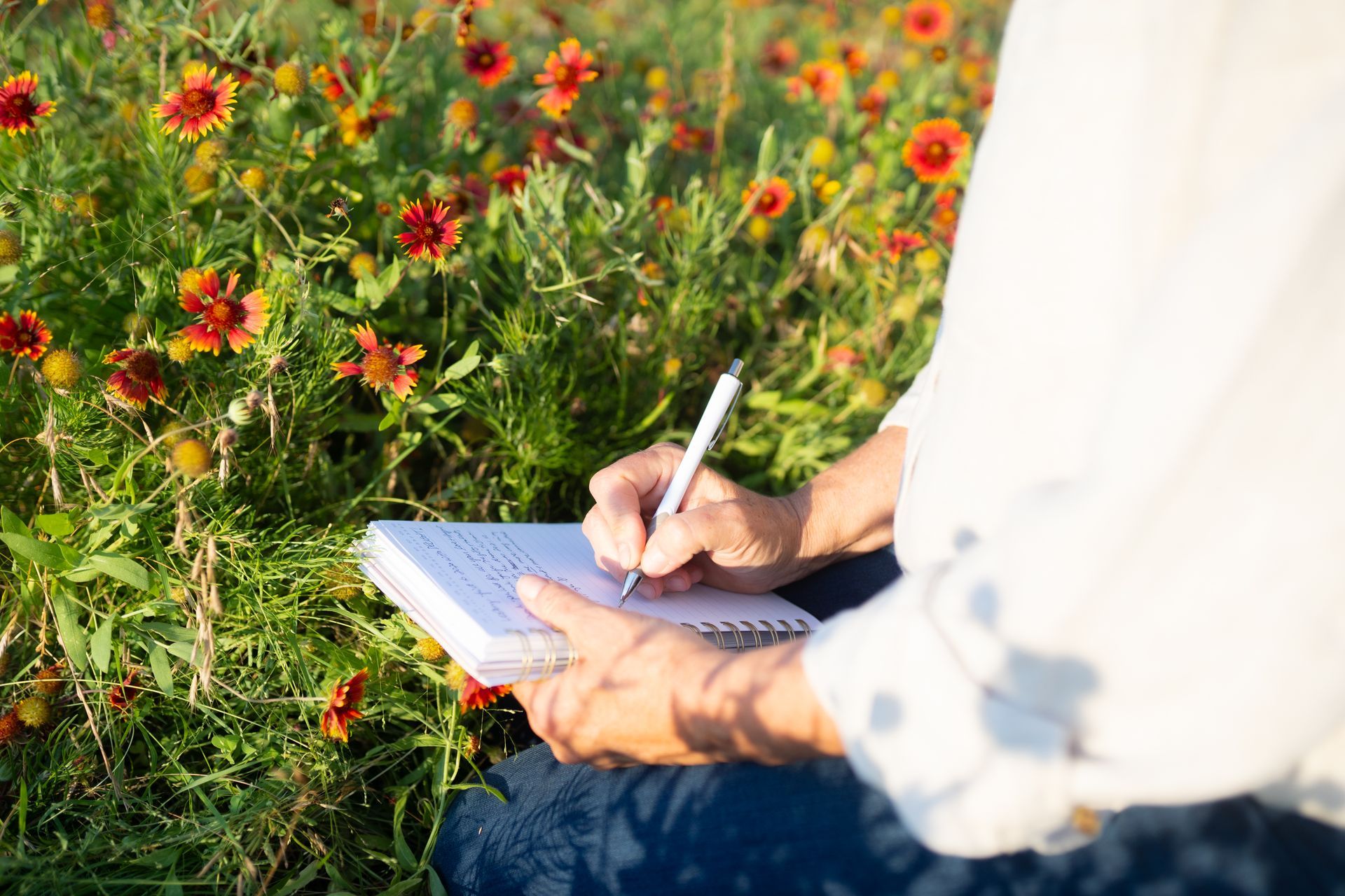 A person is sitting in a field of flowers writing in a notebook.