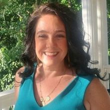 Woman with dark hair, smiling, wearing a teal top and gold necklace, outside with greenery.