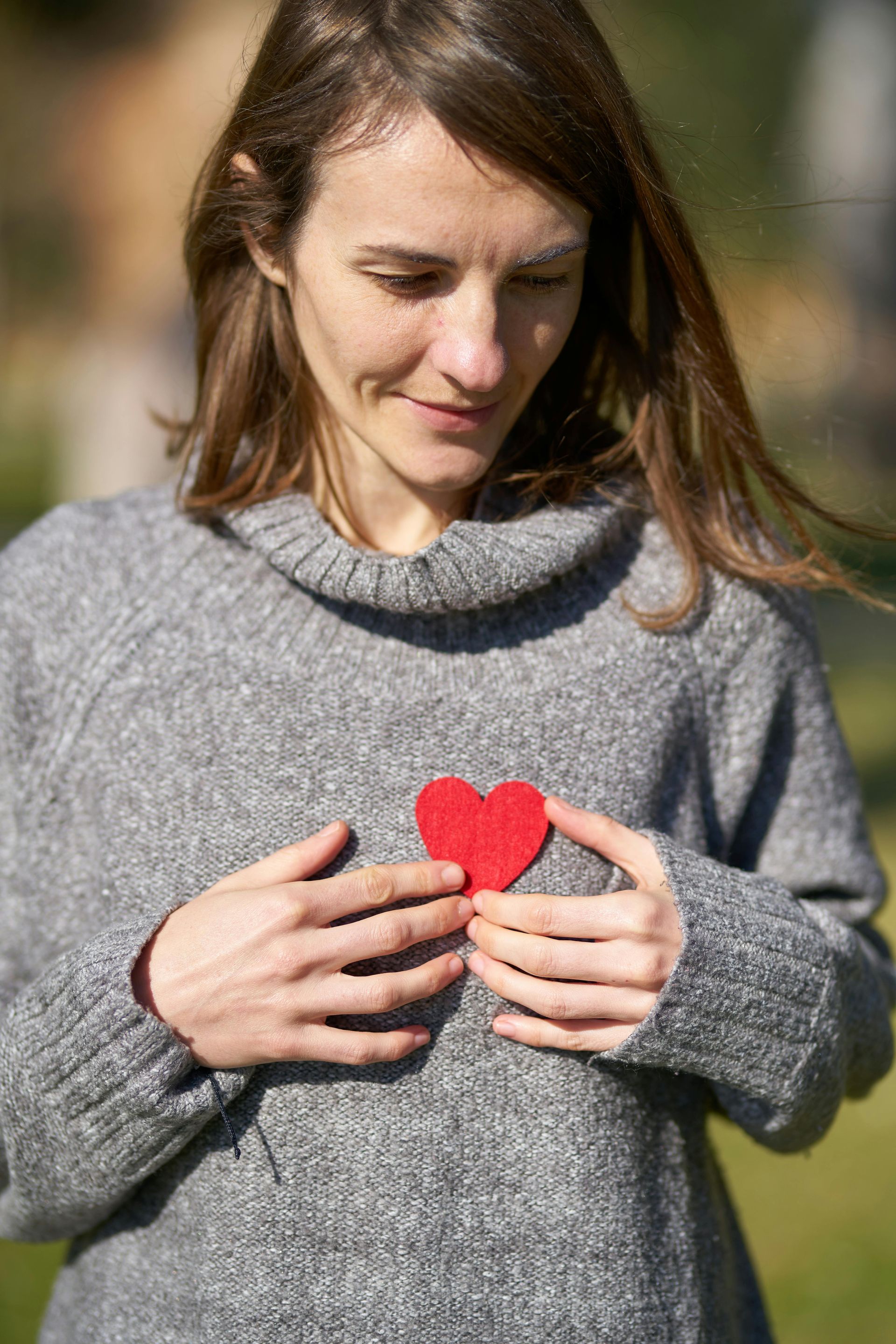 Woman holding a red heart-shaped object on her chest, wearing a gray sweater, smiling outdoors.