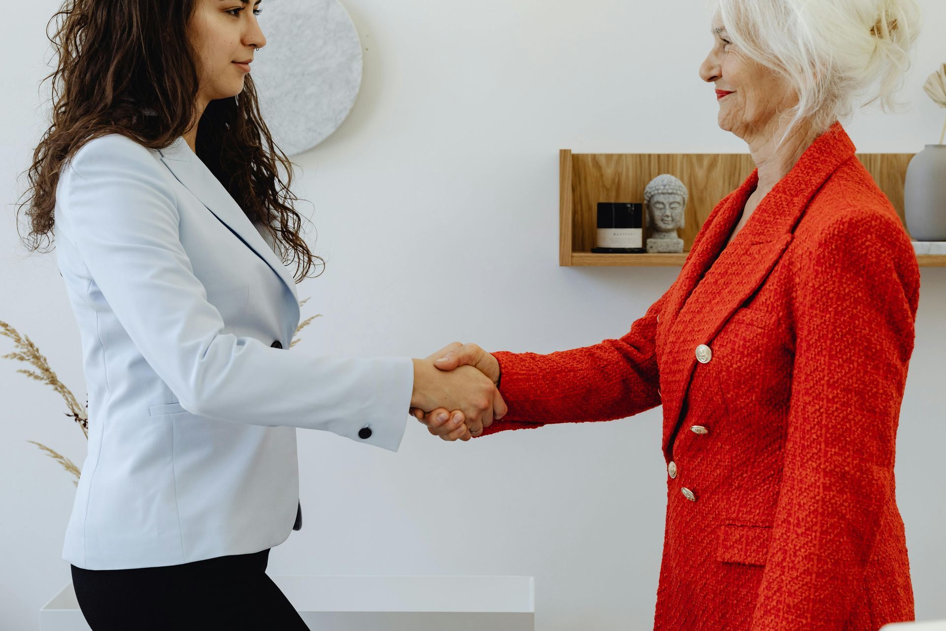 Two women in business attire shake hands in an office setting.