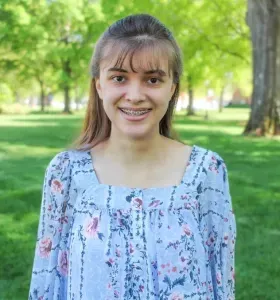A young woman wearing braces and a floral shirt is standing in a park.