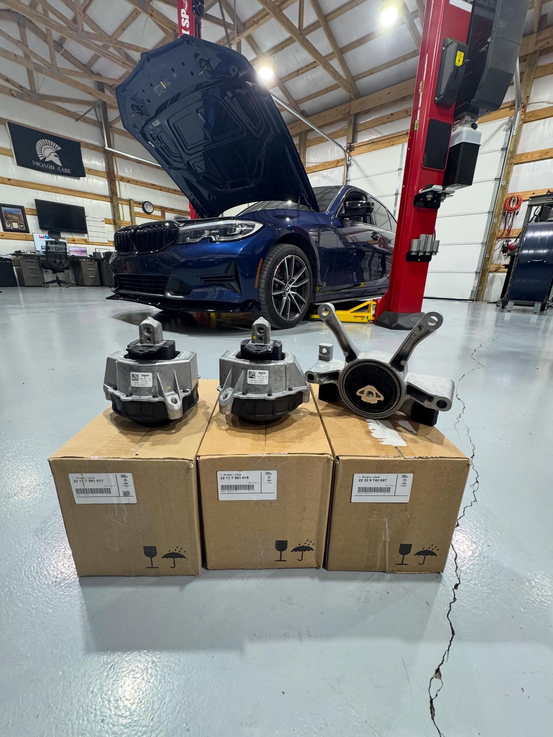 Three car parts on boxes in a workshop, with a blue car on a lift in the background | KB Motor Works