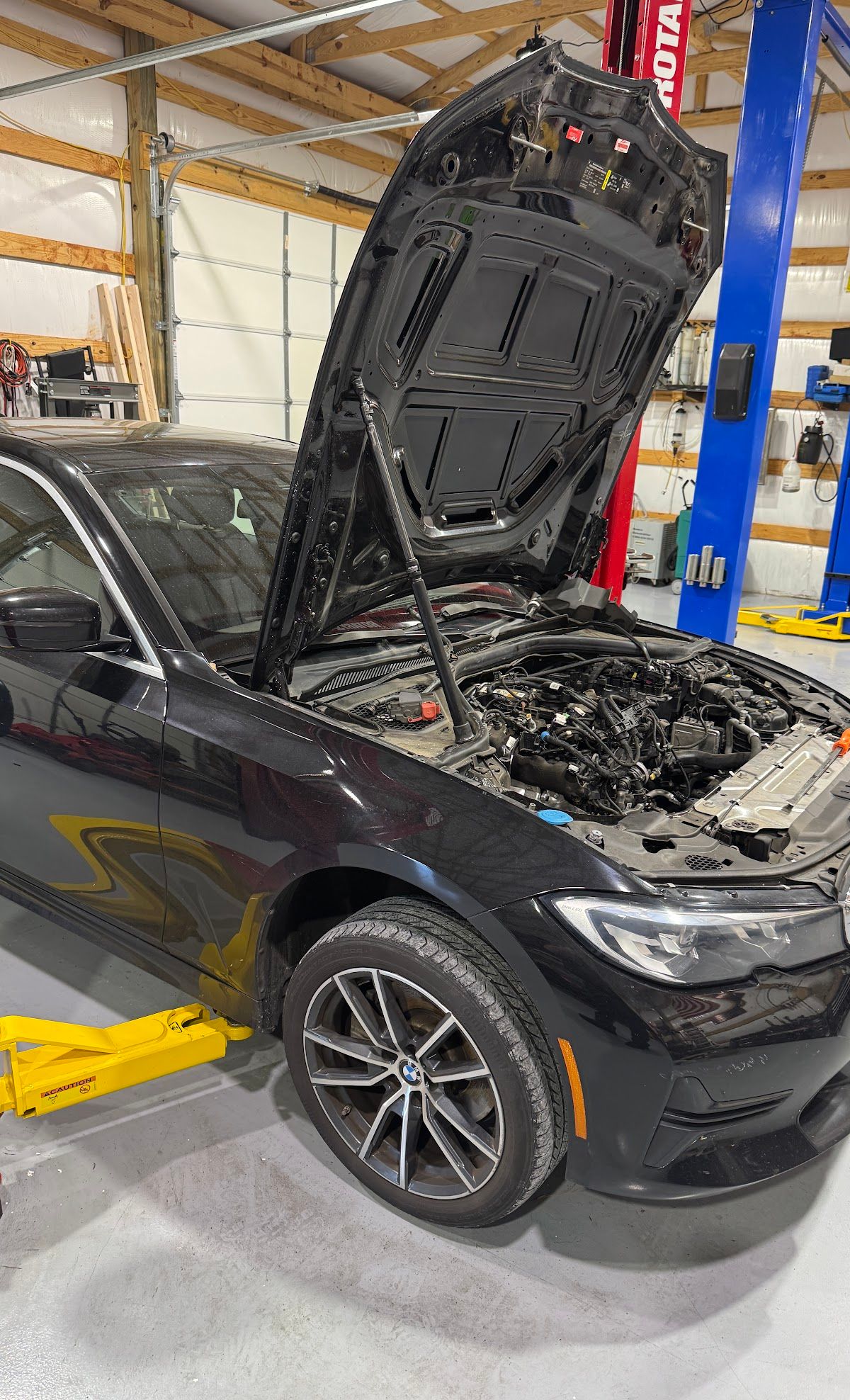 Black car with hood open in a repair shop, showing the engine bay and front wheel | KB Motor Works