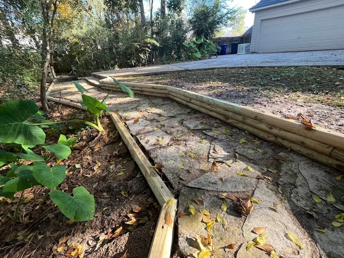A wooden path with borders winds through a yard with trees and a house in the background.