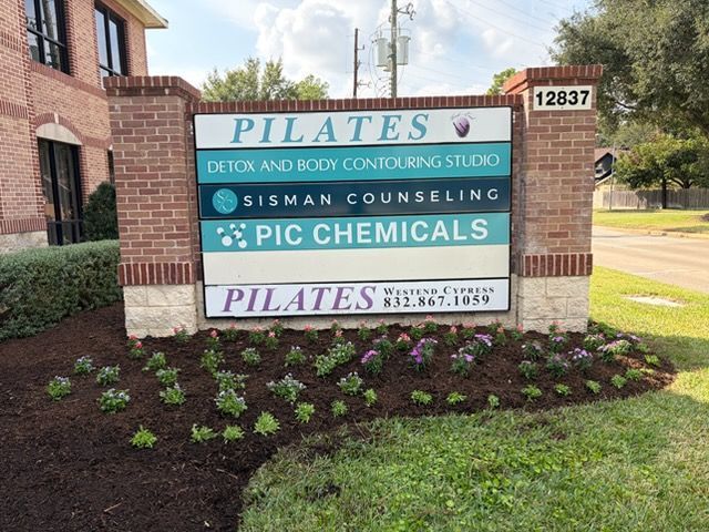 A brick business sign at 12837 listing Pilates, Sisman Counseling, and PIC Chemicals, set in a garden with small plants.