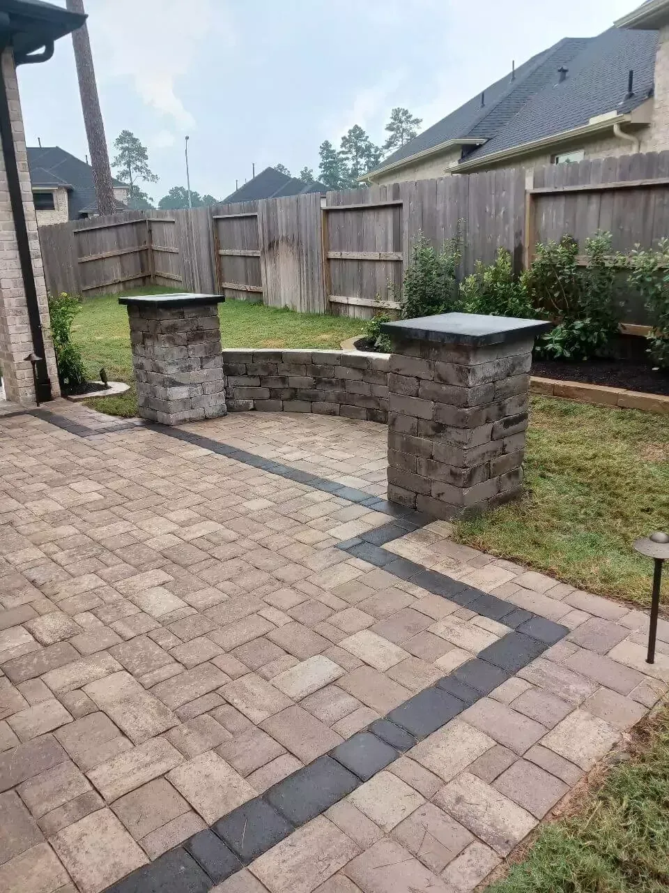 Brick patio with stone pillars and a low wall, leading to a grassy yard with a wooden fence.