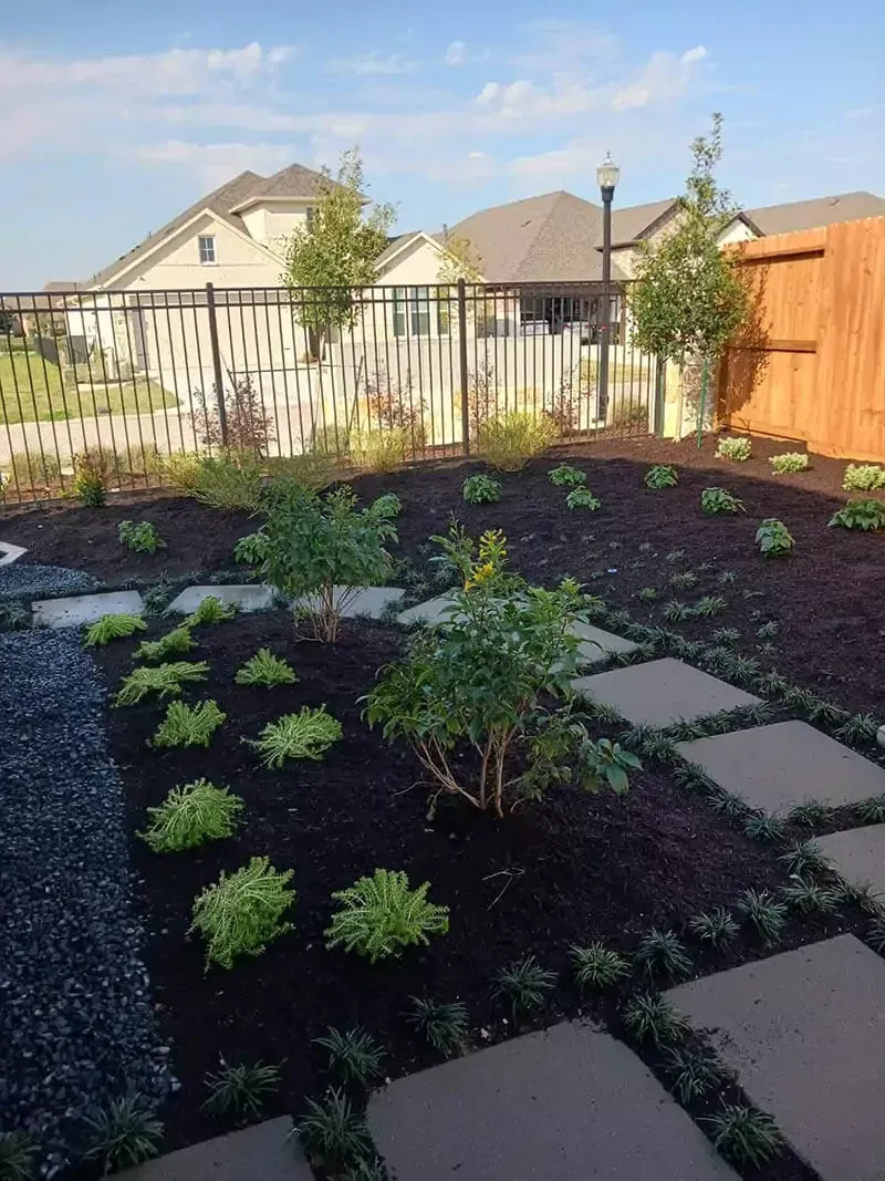 Backyard garden with stepping stones, shrubs, dark mulch, and a fence.