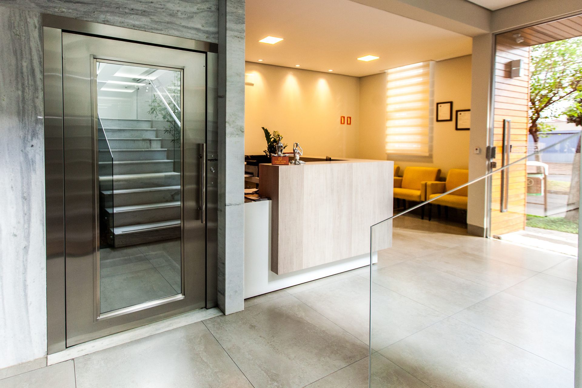 Hall de entrada moderno com elevador de vidro, balcão de recepção em madeira clara, cadeiras amarelas e piso de mármore.