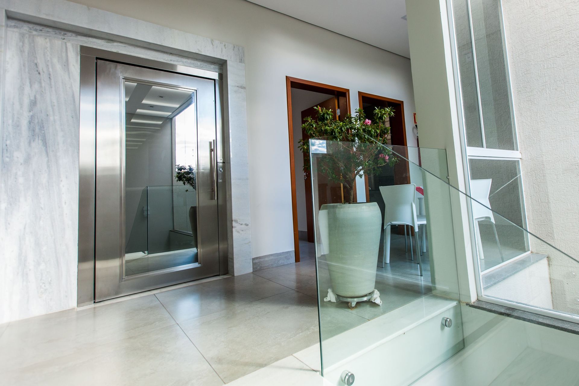 Porta de elevador em aço inoxidável em frente à um vaso de planta em um patamar luminoso e minimalista com piso branco.