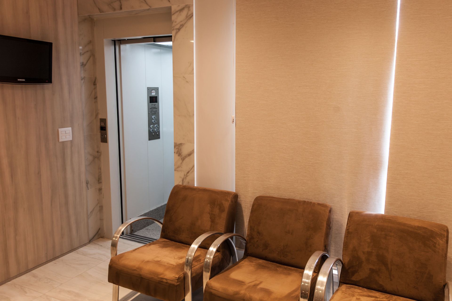 Área de espera com três cadeiras marrons de frente para um elevador aberto, em uma sala com paredes e piso claros.