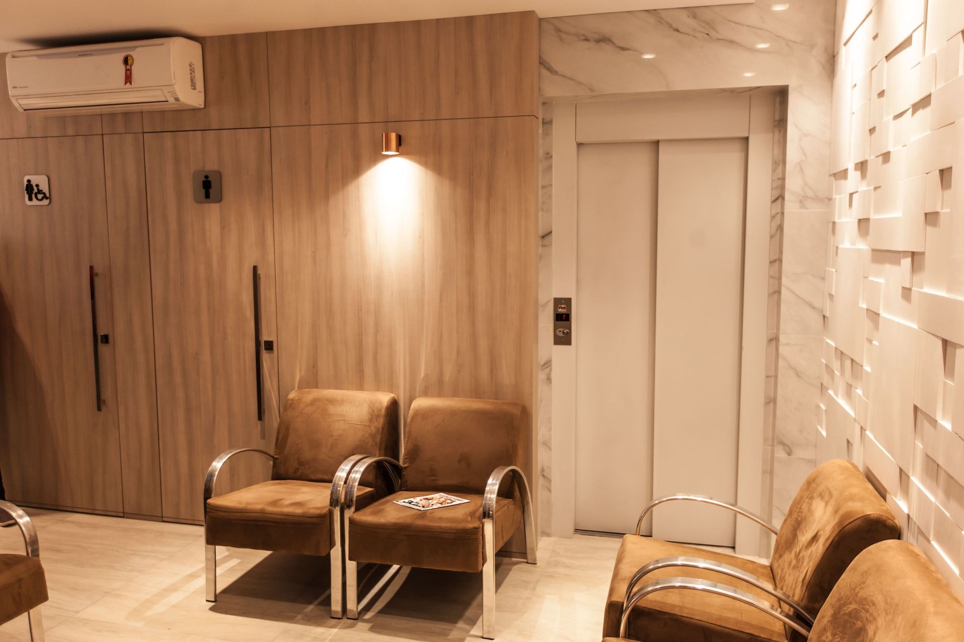Sala de espera com quatro cadeiras marrons, uma parede revestida de madeira com ar-condicionado e uma porta de elevador.