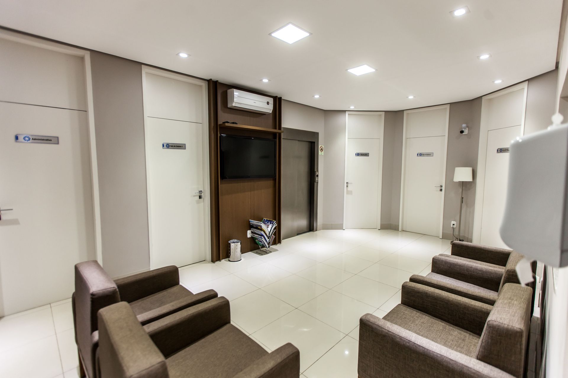 Sala de espera com cadeiras marrons, portas brancas, uma TV fixada na parede e um elevador em um espaço claro e iluminado.