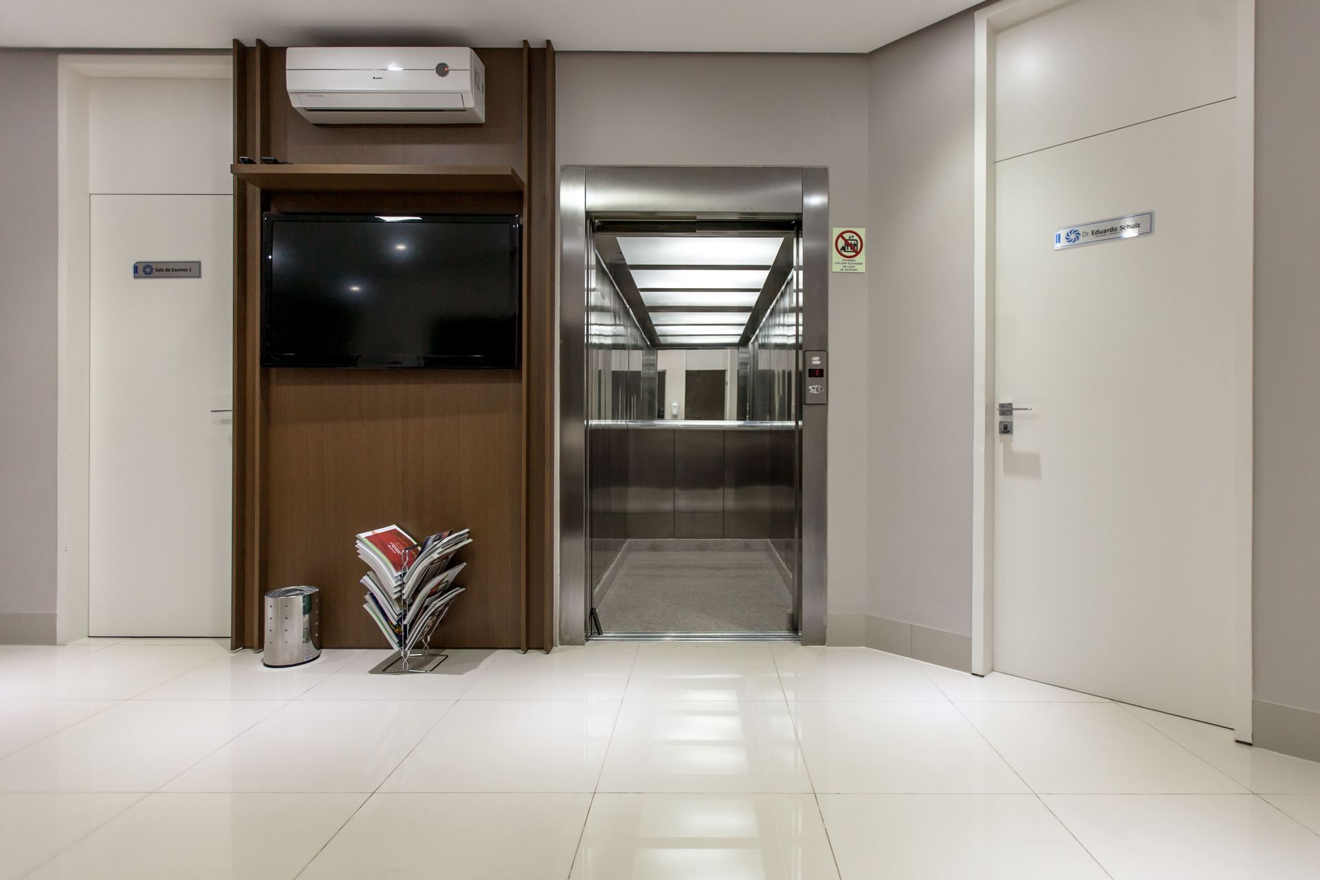 Hall de entrada moderno com uma TV na parede revestida de madeira, um elevador aberto e portas brancas em ambos os lados.