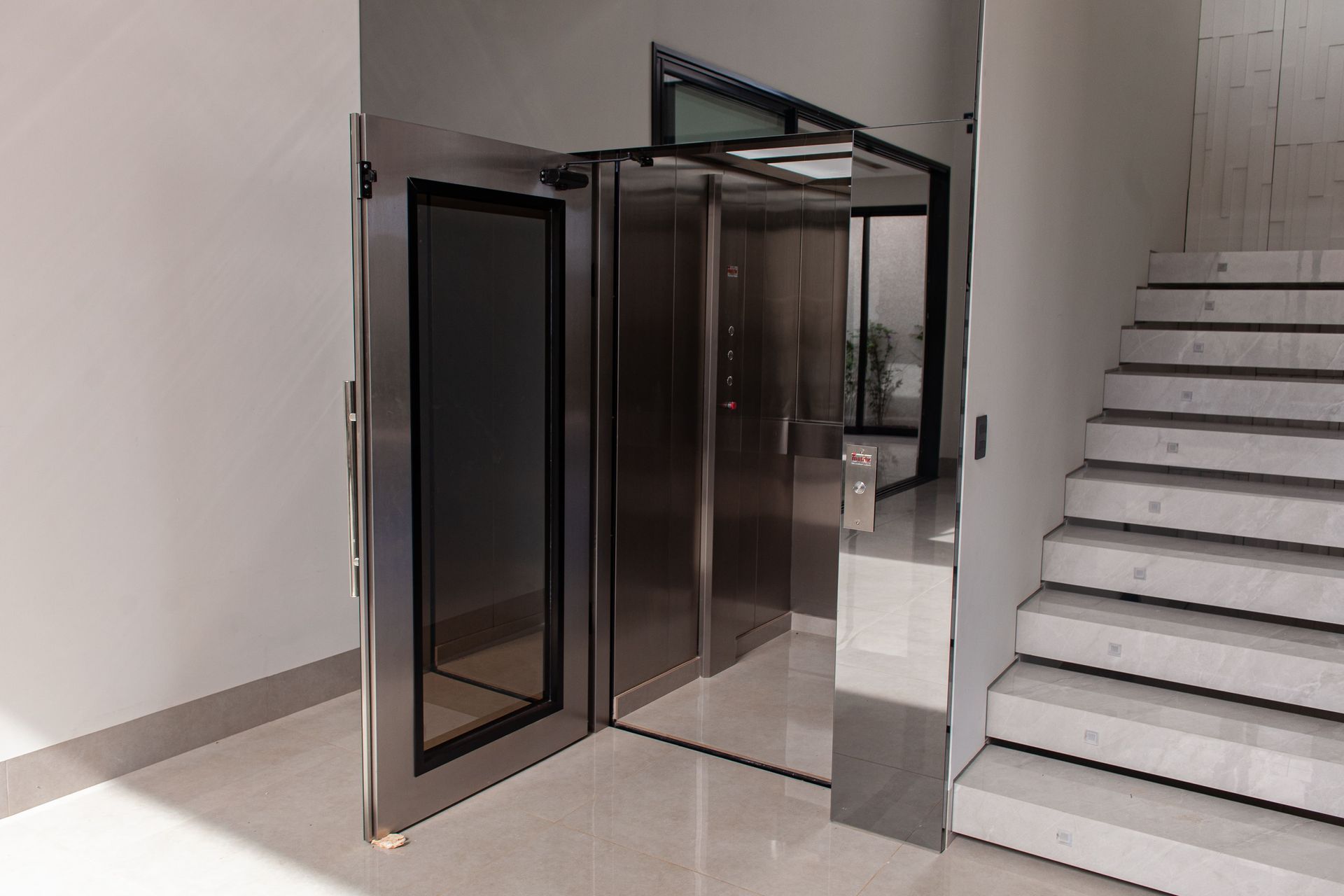 Elevador moderno em aço inoxidável com porta de vidro aberta, posicionado ao lado de uma escada minimalista de concreto.