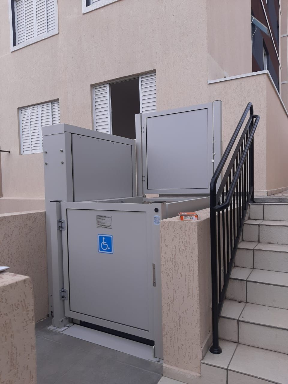 Plataforma elevatória vertical cinza para cadeirante de rodas, instalada ao lado de uma escada de concreto de um edifício.