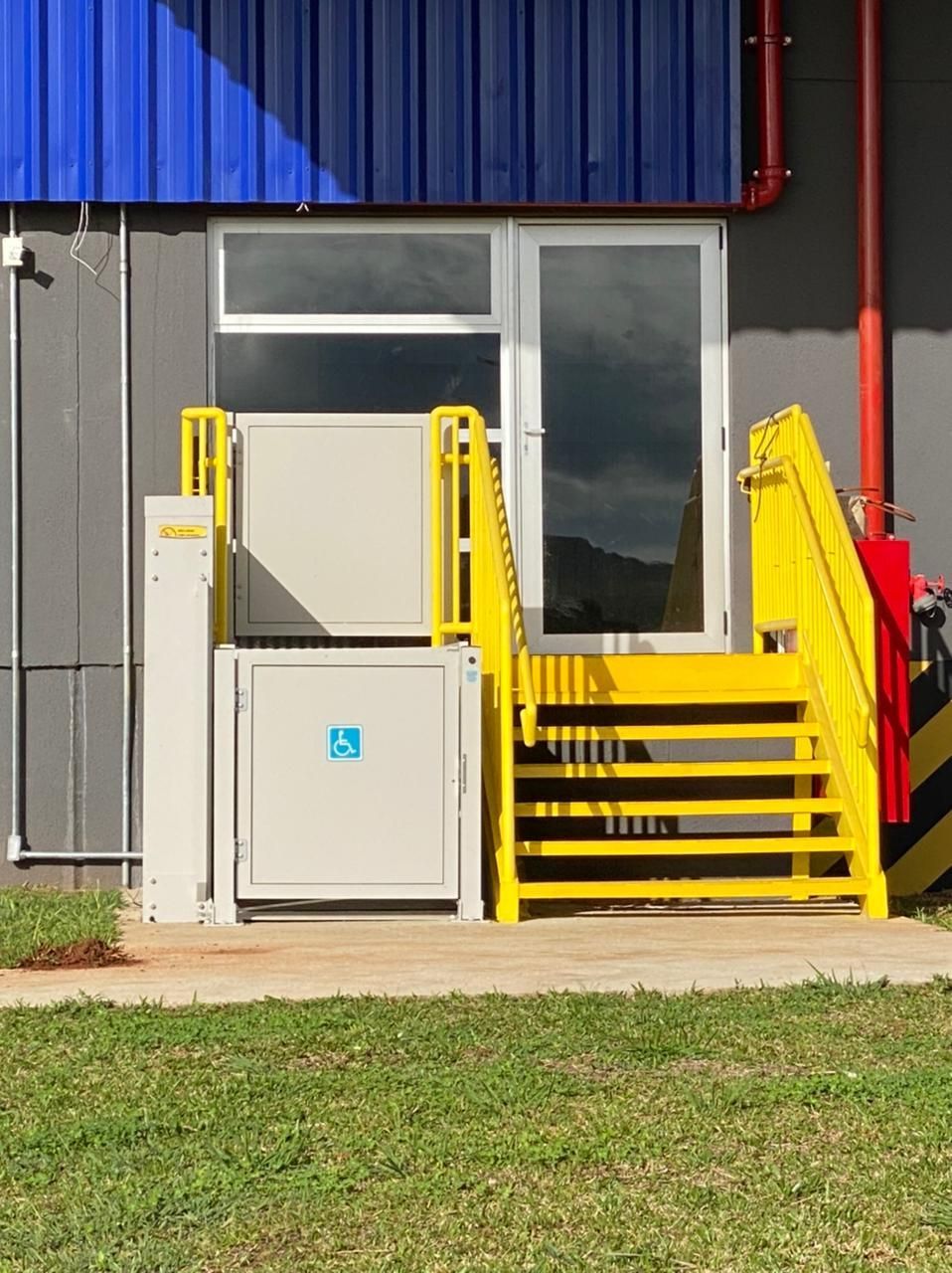 Escada metálica amarela com um elevador para cadeiras de rodas, na entrada de um prédio com parede cinza e revestimento azul.