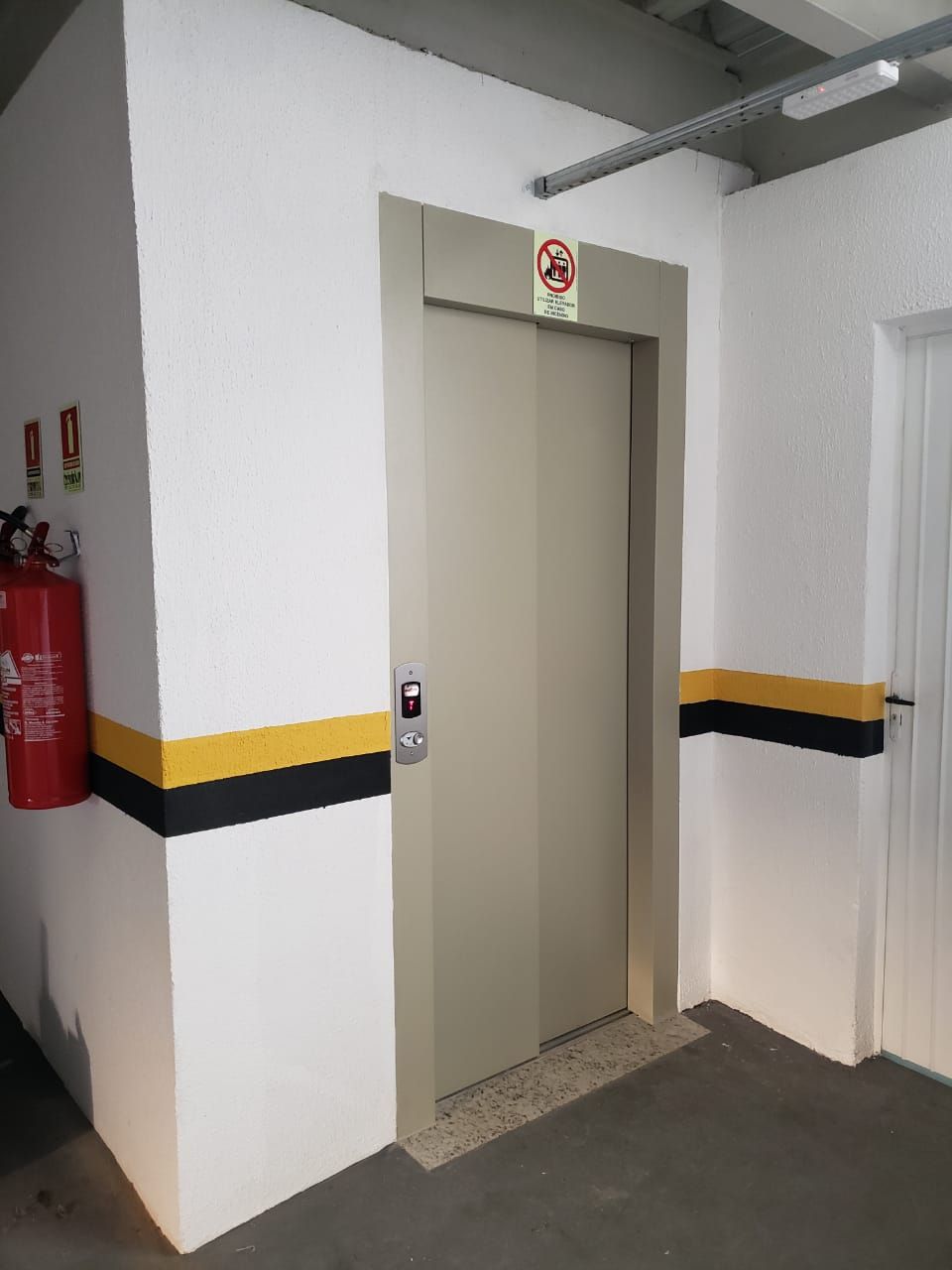 Elevador numa garagem de estacionamento com portas bege, paredes brancas e faixas horizontais de segurança amarelas e pretas.